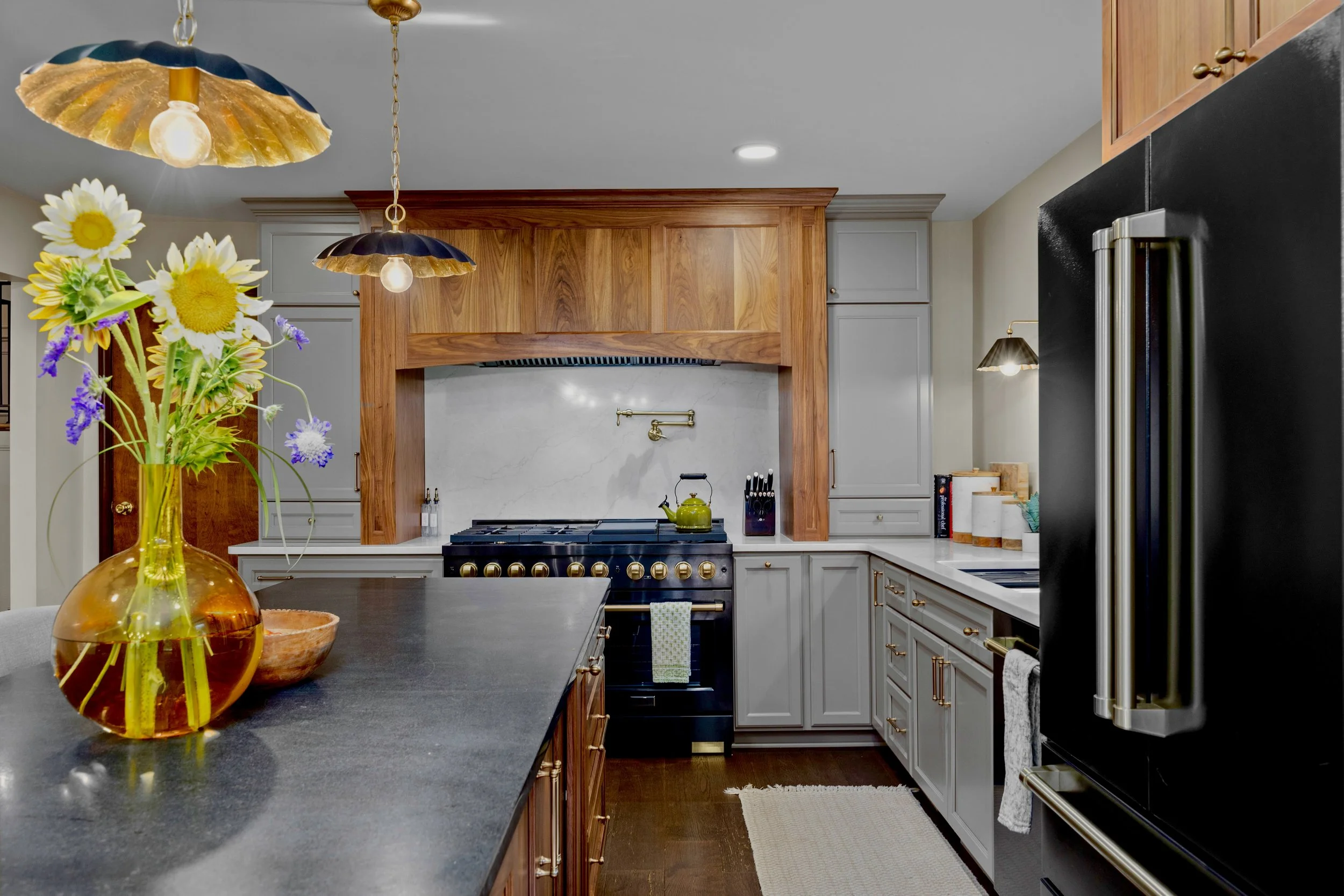 A cozy kitchen with gray cabinets, a black stove with gold knobs, wooden accents, hanging pendant lights, and a decorative vase with sunflowers and purple flowers on a dark countertop.
