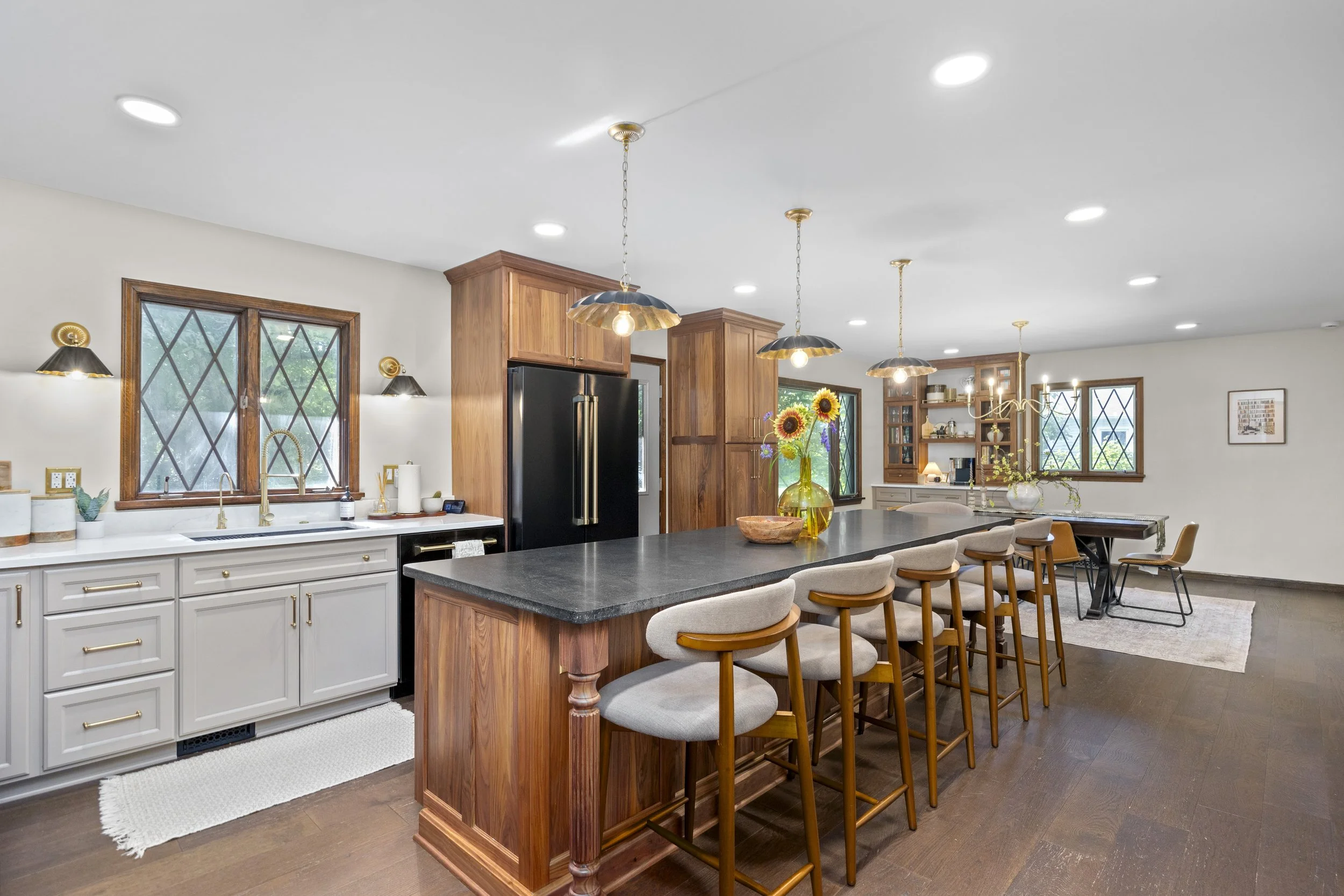 Open-concept kitchen and dining area with white cabinetry, black countertops, wooden accents, and large windows with diamond-shaped panes; pendant and wall lighting, fresh flower arrangements, and hardwood floors.
