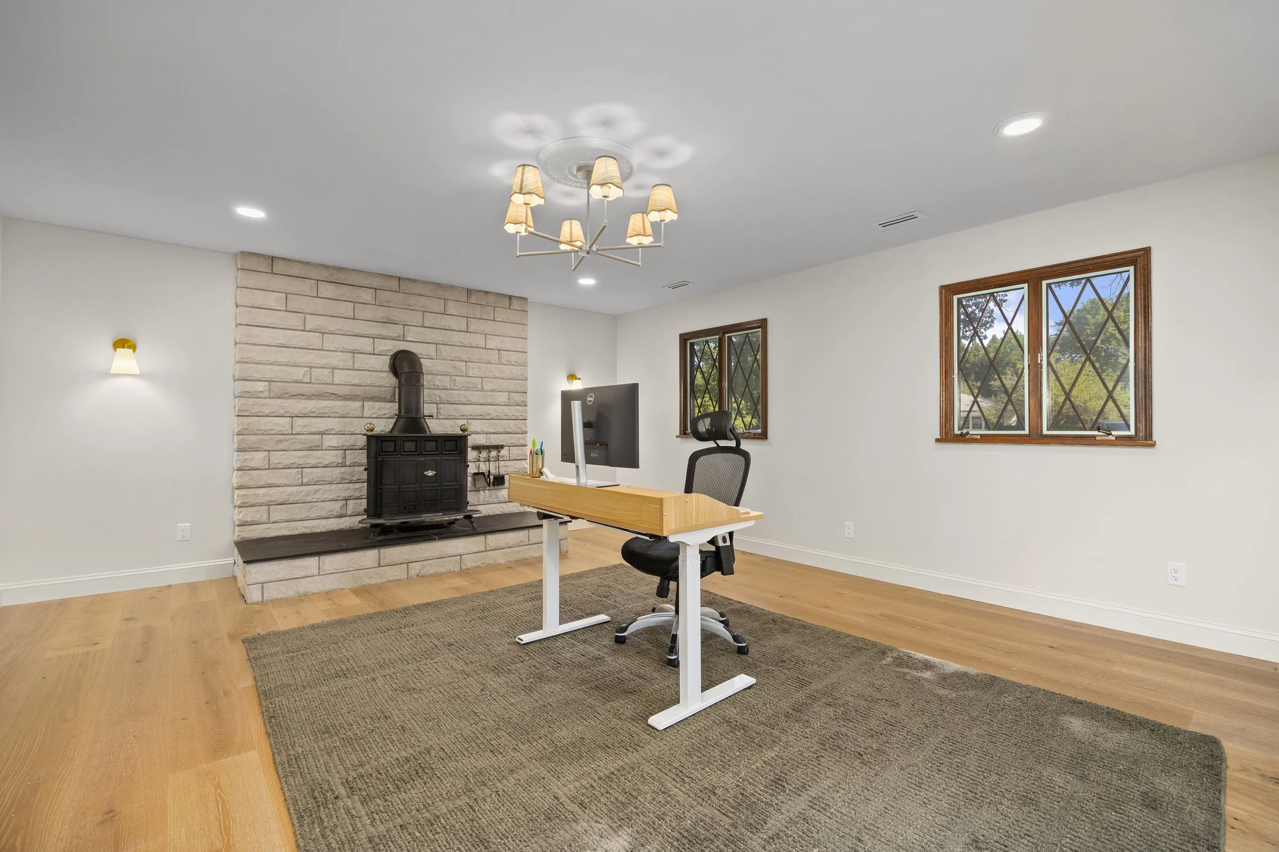Bright office space with white walls, wood flooring, two windows, a stone fireplace with a black stove, modern chandelier with beige lampshades, wall sconces, a desk with a computer, office chair, and a rug.