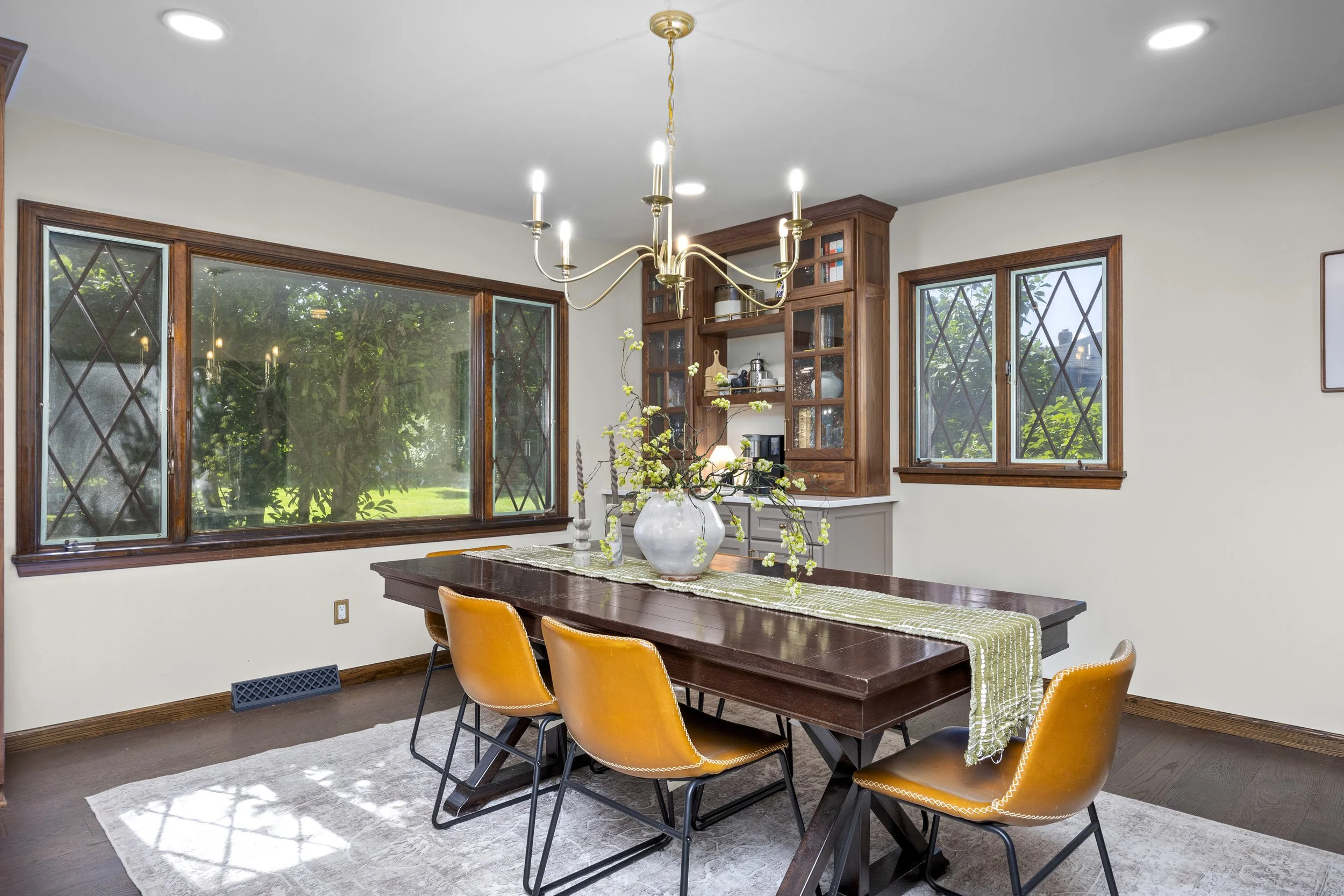 A dining room with a dark wooden table and six orange chairs with black legs. A white vase with branches and flowers is on the table, which has a green and white striped table runner. The room has three large windows with wooden frames and diamond-sh