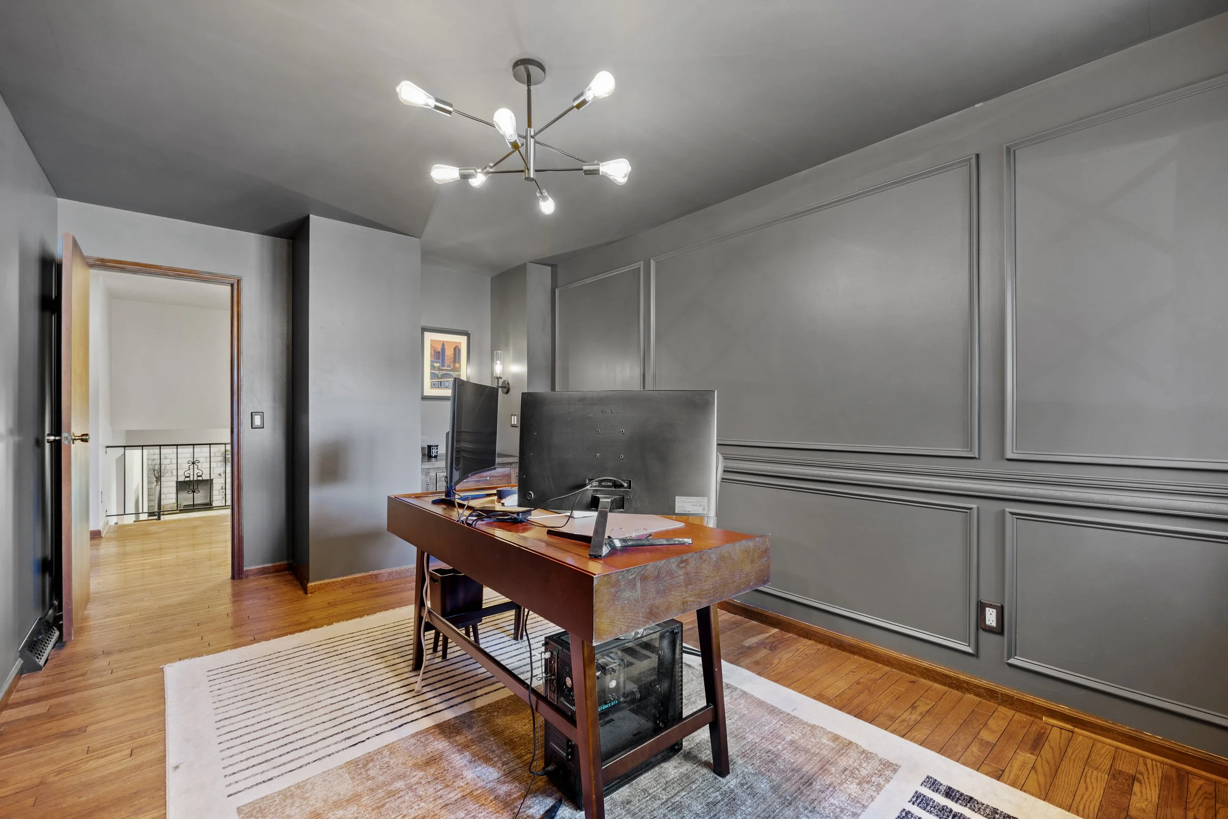 A small office space with a wooden desk, computer monitor, and a computer tower underneath. The room has gray walls with decorative molding, wooden flooring, and a white rug. There is a modern chandelier with multiple exposed bulbs hanging from the c