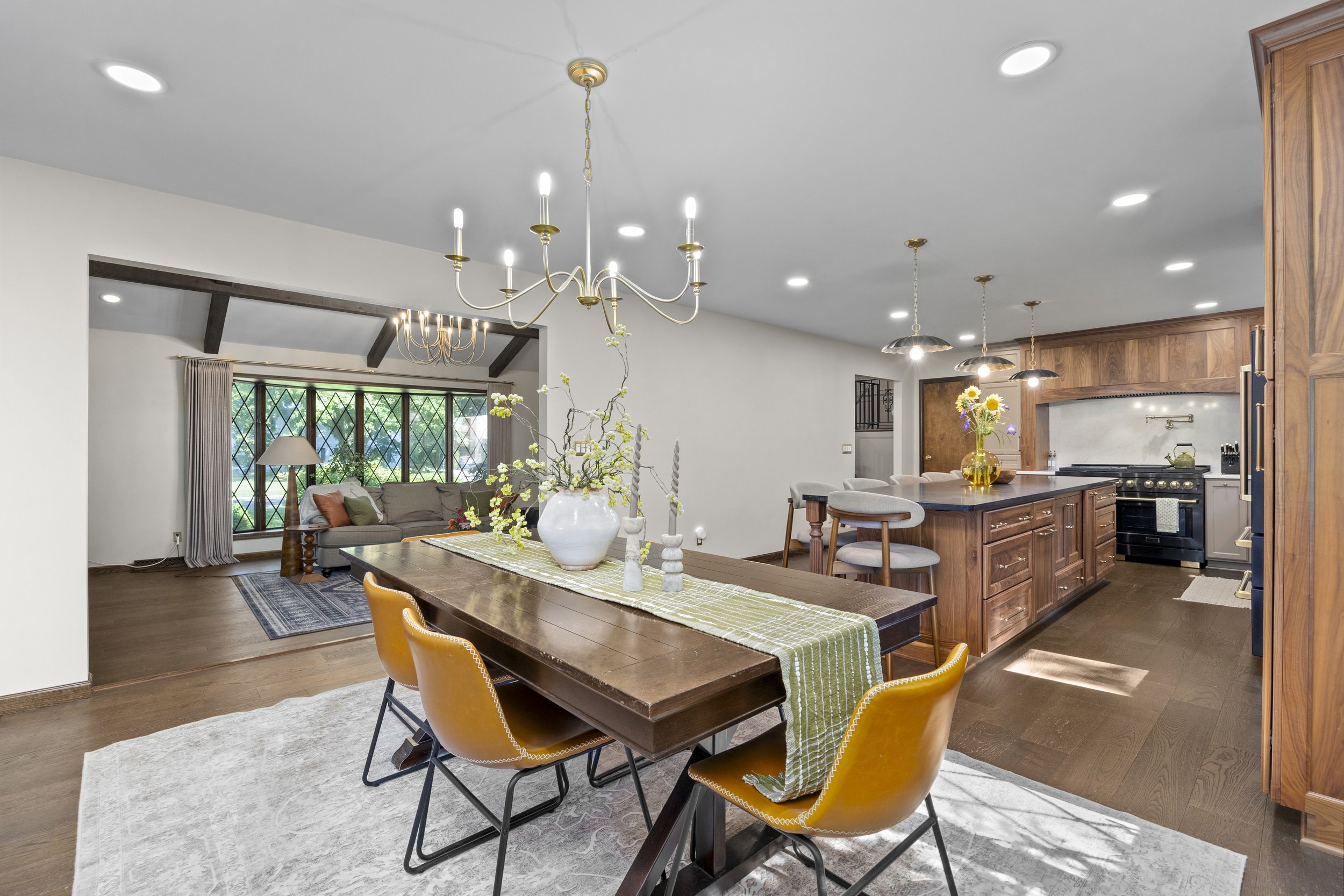 Open-concept living and dining area with hardwood flooring, a wooden dining table with yellow chairs, a kitchen with wooden cabinets, black stove, and a breakfast bar. Bright lighting and large windows with curtains provide natural light.