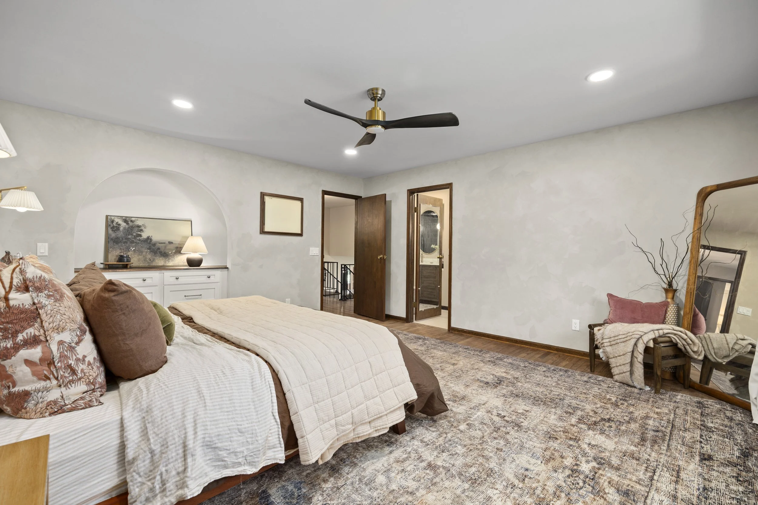 A cozy bedroom with a bed covered in white and brown bedding, decorative pillows, a side table with a lamp, a rug, a wooden chair with a blanket, and an open door leading to a bathroom.