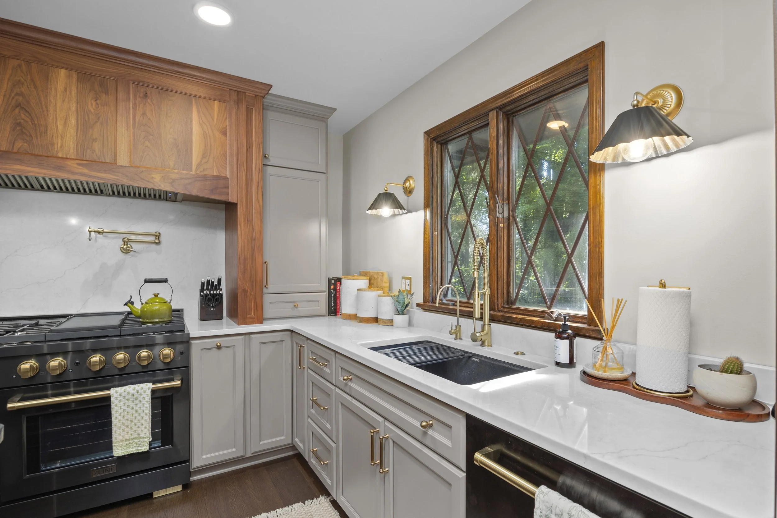 A modern kitchen with white countertops, gray cabinets, wooden window frames, a black stove, and brass accents. There are decorative plants, kitchen utensils, and a paper towel roll on the counter.