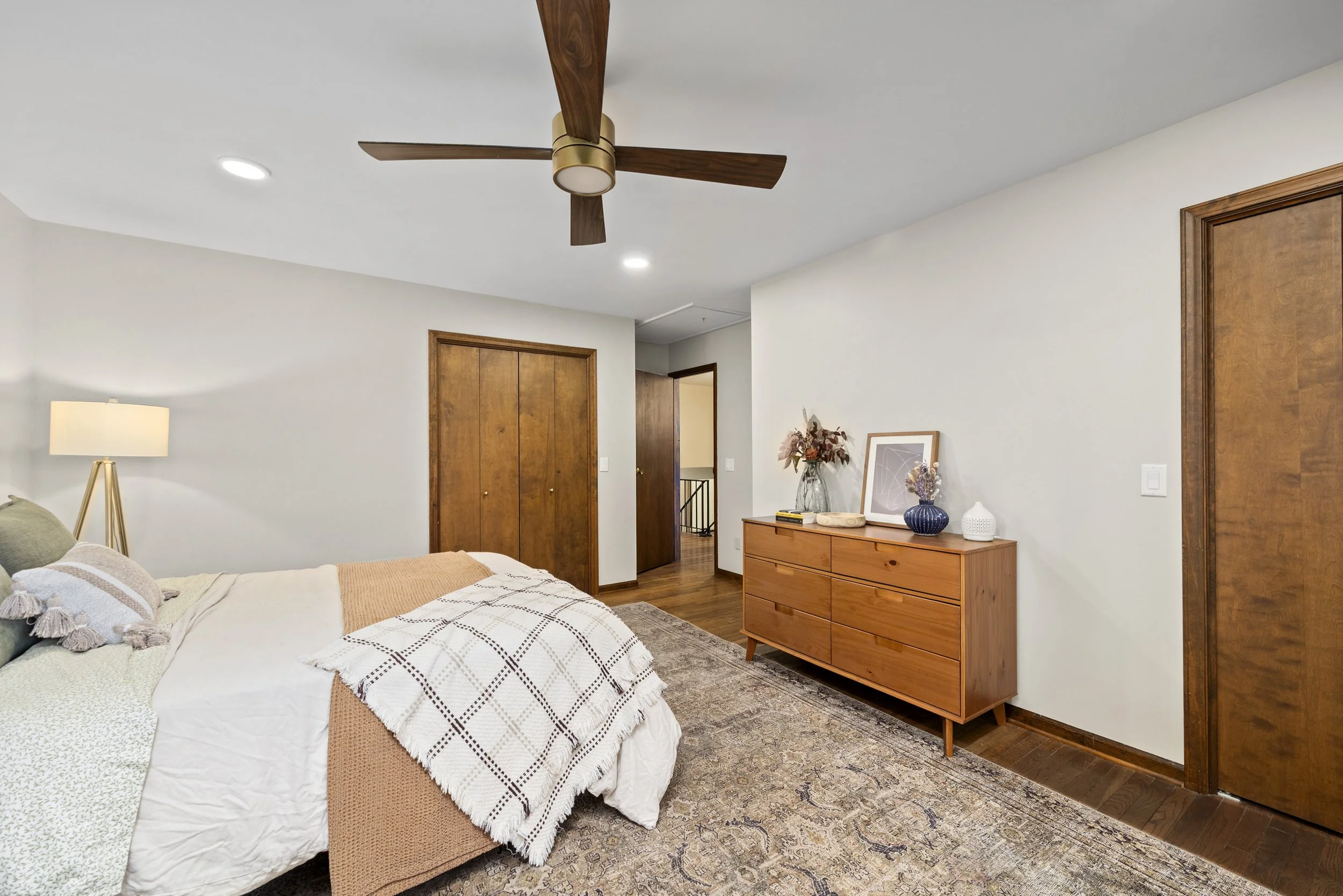 Room with a bed, nightstand, dresser, and decorative accessories, featuring a ceiling fan and wooden doors.