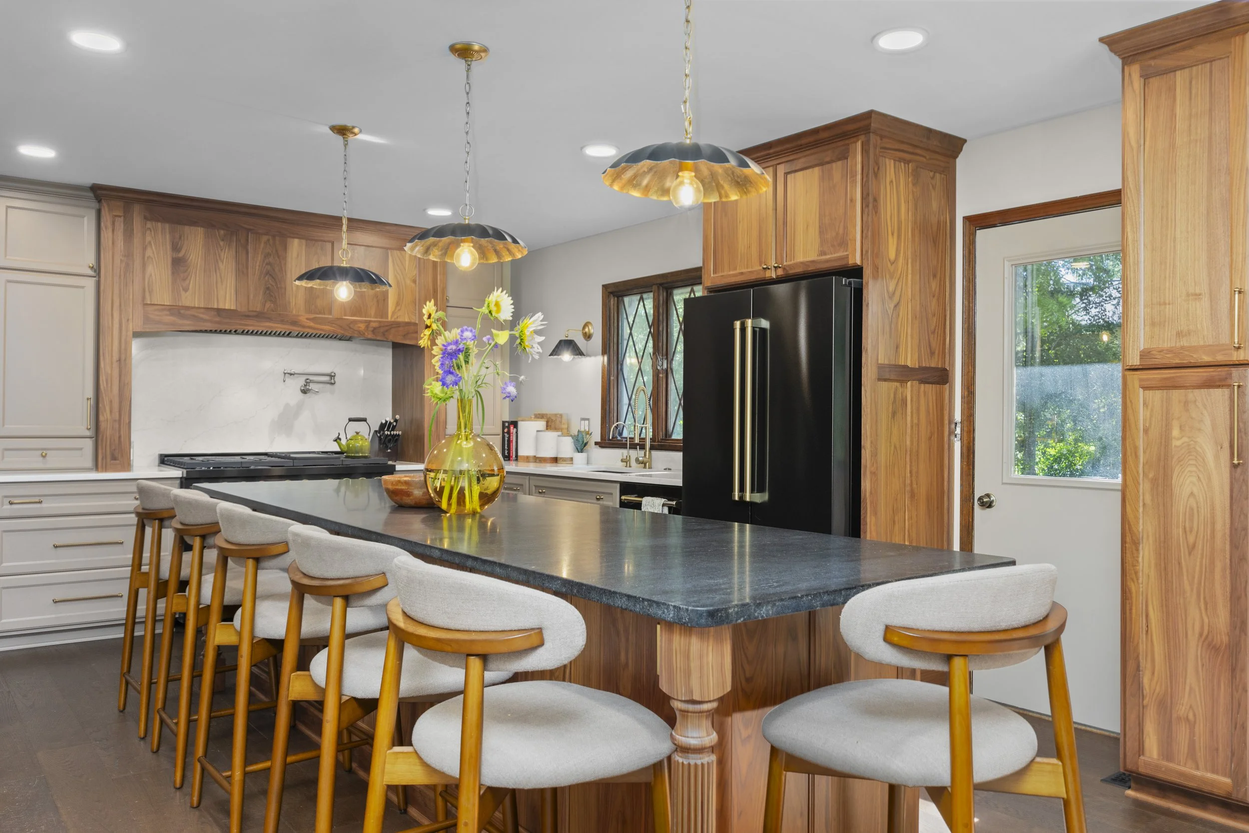 A spacious kitchen with a black island countertop, wooden cabinets, and a vase of colorful flowers on the island. There are four cream-colored stools with wooden legs placed at the island, and pendant lights hanging from the ceiling. The kitchen has 