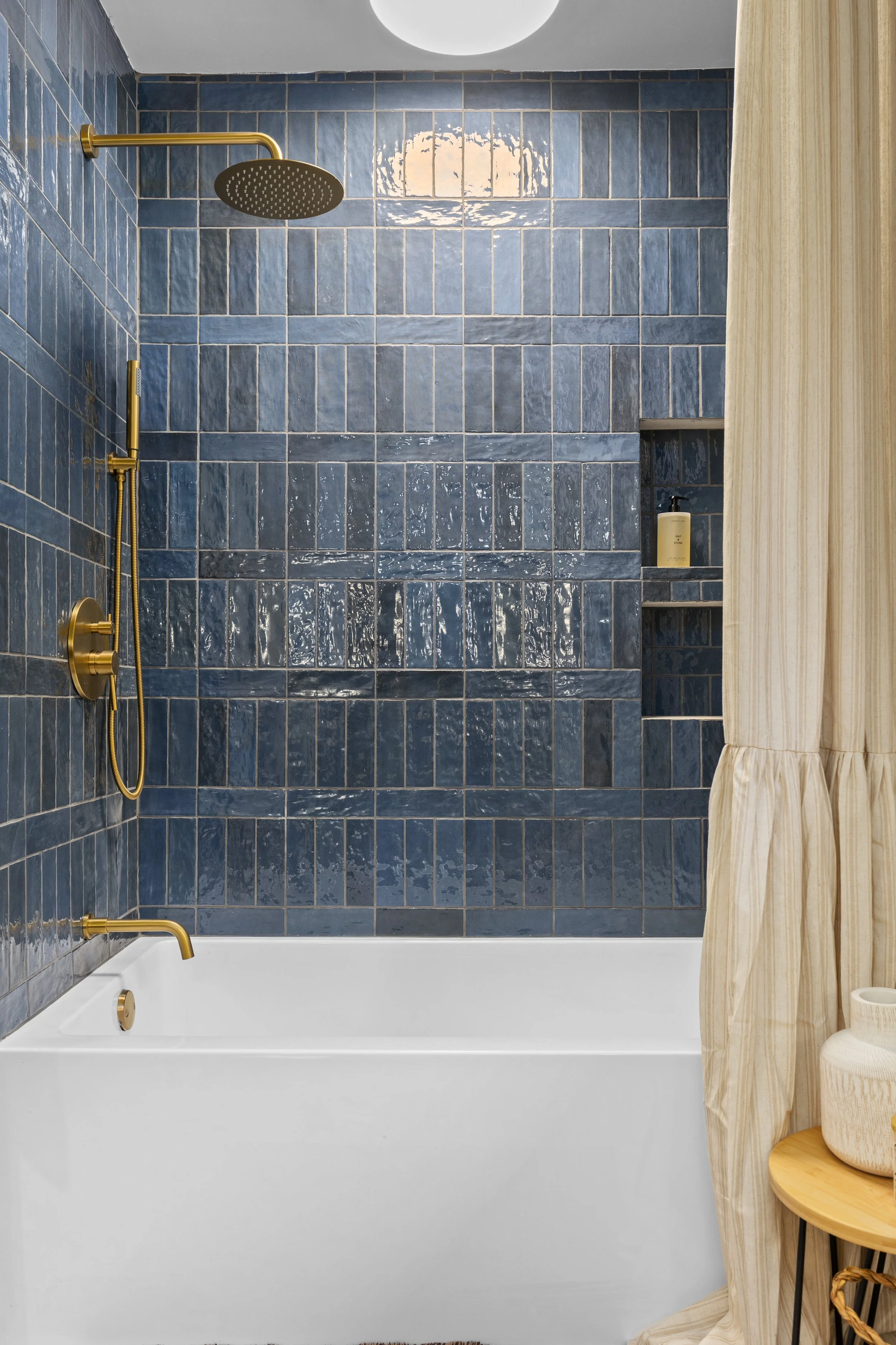 Bathroom with navy blue tiled walls, gold fixtures including a showerhead, faucet, and control knob, a built-in shelf with a bottle of body lotion, a white bathtub with gold drain and faucet, beige curtain with striped pattern, and a round wooden sid