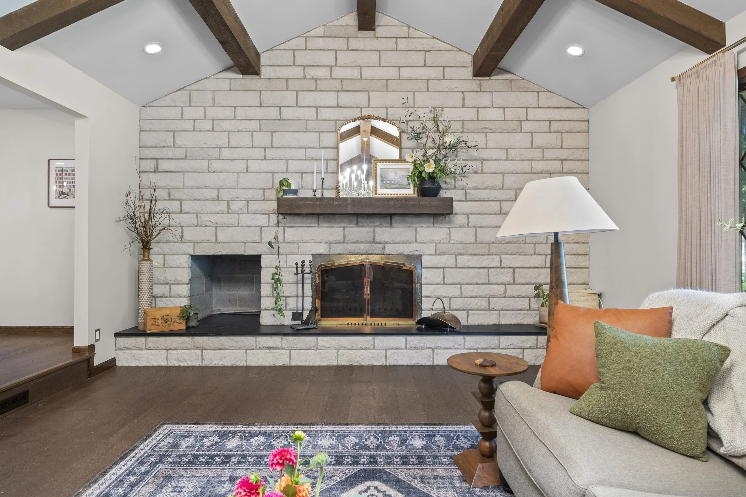 Living room with white brick fireplace, wooden beams, decorative mirror, plants, and a comfortable sofa with colorful pillows.