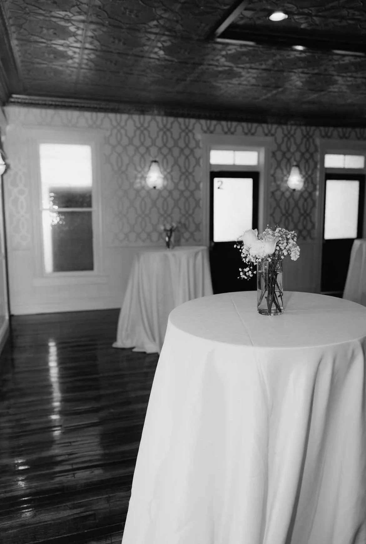 A black and white photo of an elegantly decorated interior room with multiple tall windows, wallpaper with a geometric pattern, and a ceiling with an intricate wood design. There are standing tables with white tablecloths, one with a small floral arrangement in a glass vase. The room appears to be set up for an event or gathering.