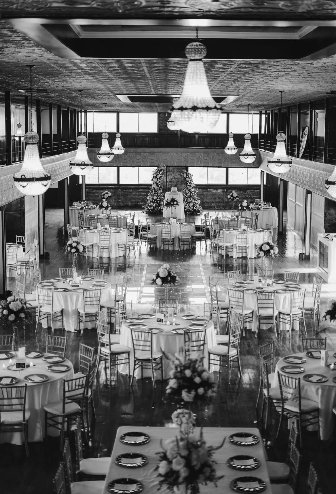 Black and white photo of an elegant banquet hall decorated for a wedding reception with round tables, flower centerpieces, chandeliers, and large windows.