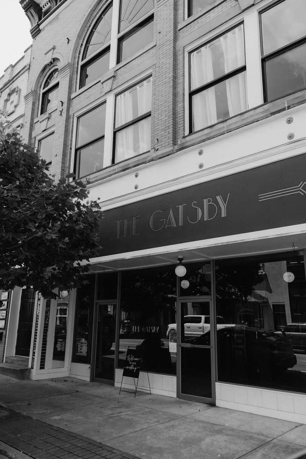 Black and white photo of a storefront with a sign that reads 'The Gatsby'. The building has large windows, ornate brickwork, and decorative elements. There is a small chalkboard sign outside the entrance.
