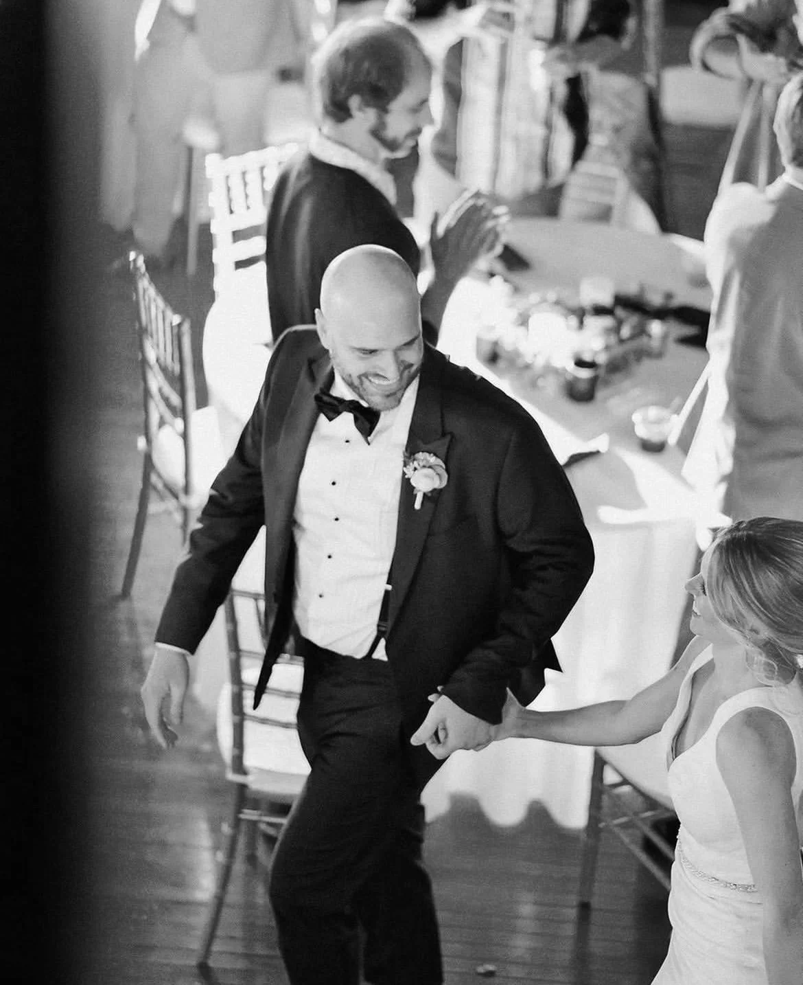 Black and white photo of a groom in a tuxedo holding hands with a bride in a wedding dress during a wedding reception.