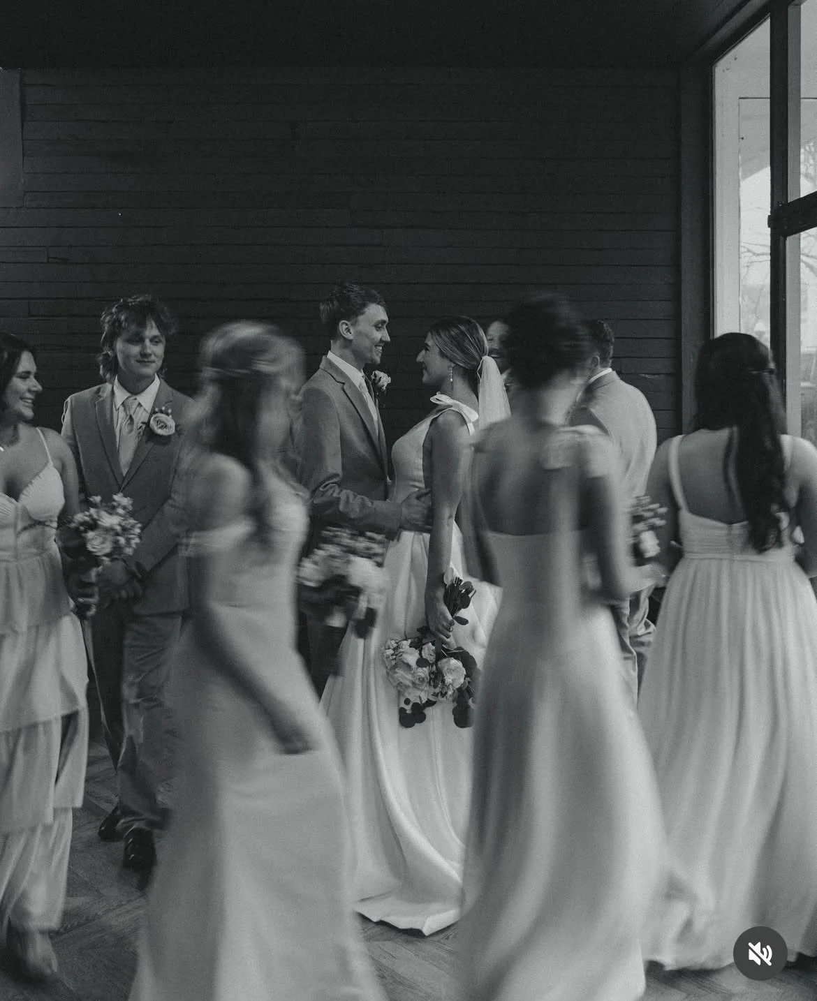 A black-and-white photo of a wedding ceremony with the bride and groom standing face to face, surrounded by bridesmaids and groomsmen, some holding bouquets, in an indoor venue with wooden walls and a large window.