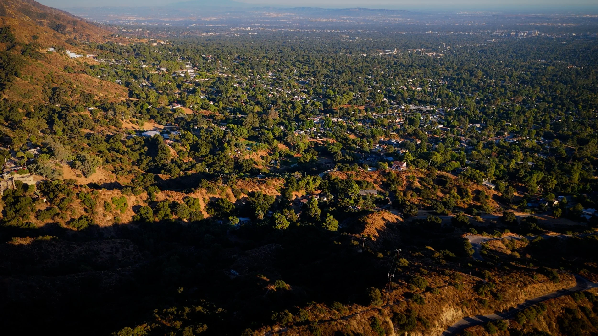 Aerial view of a mountainous suburban area with many houses surrounded by green trees and vegetation, with a distant cityscape in the background.