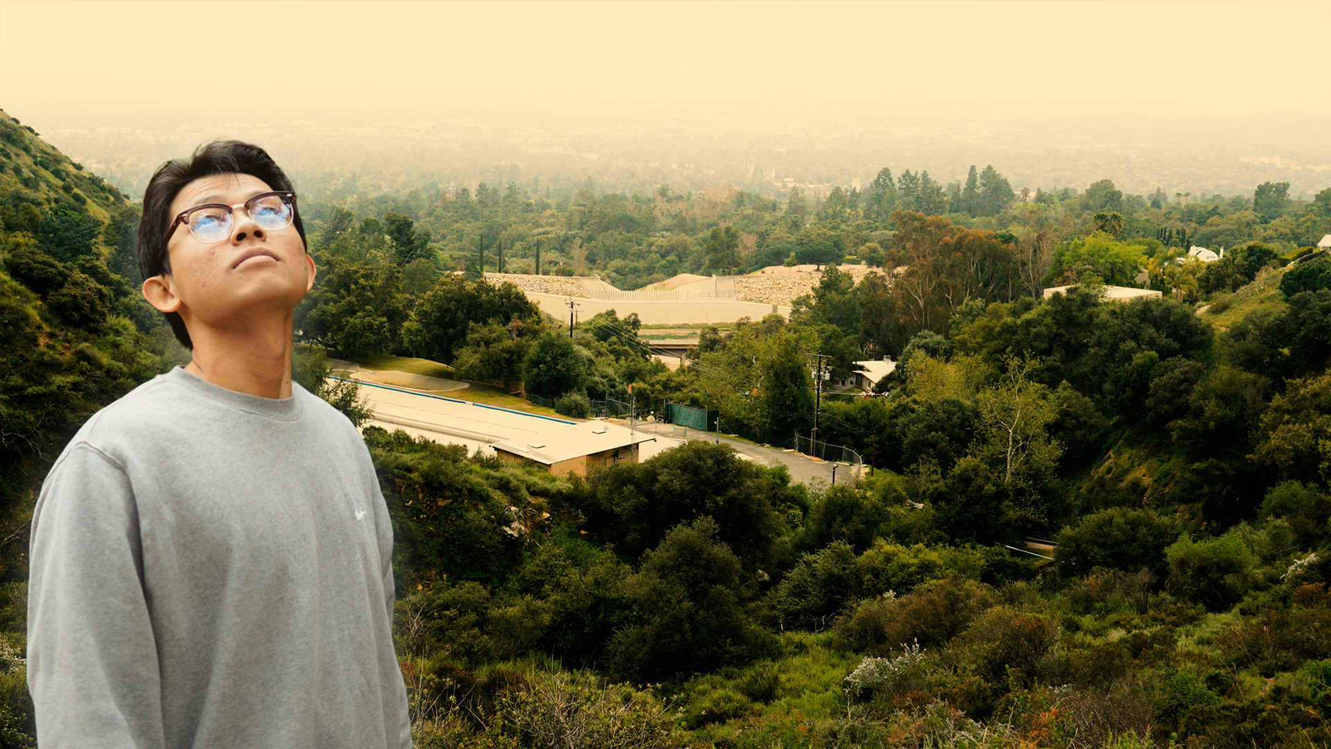 A man wearing glasses and a gray Nike sweatshirt looking upward, with a lush green hillside and distant cityscape in the background located in Altadena California