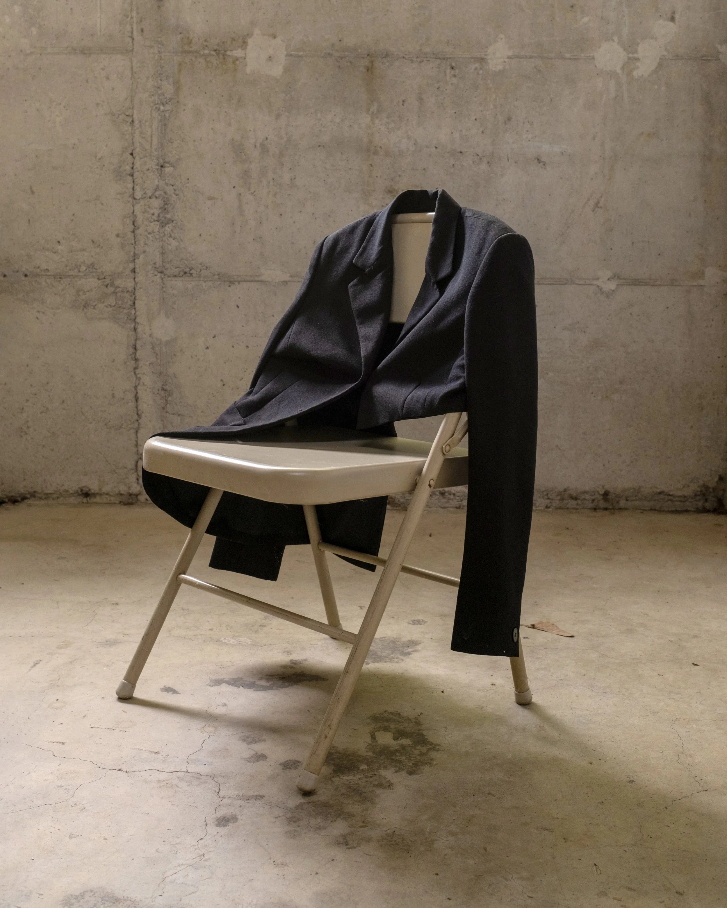 Silla blanca con chaqueta oscura colgada, en un fondo de pared de cemento.