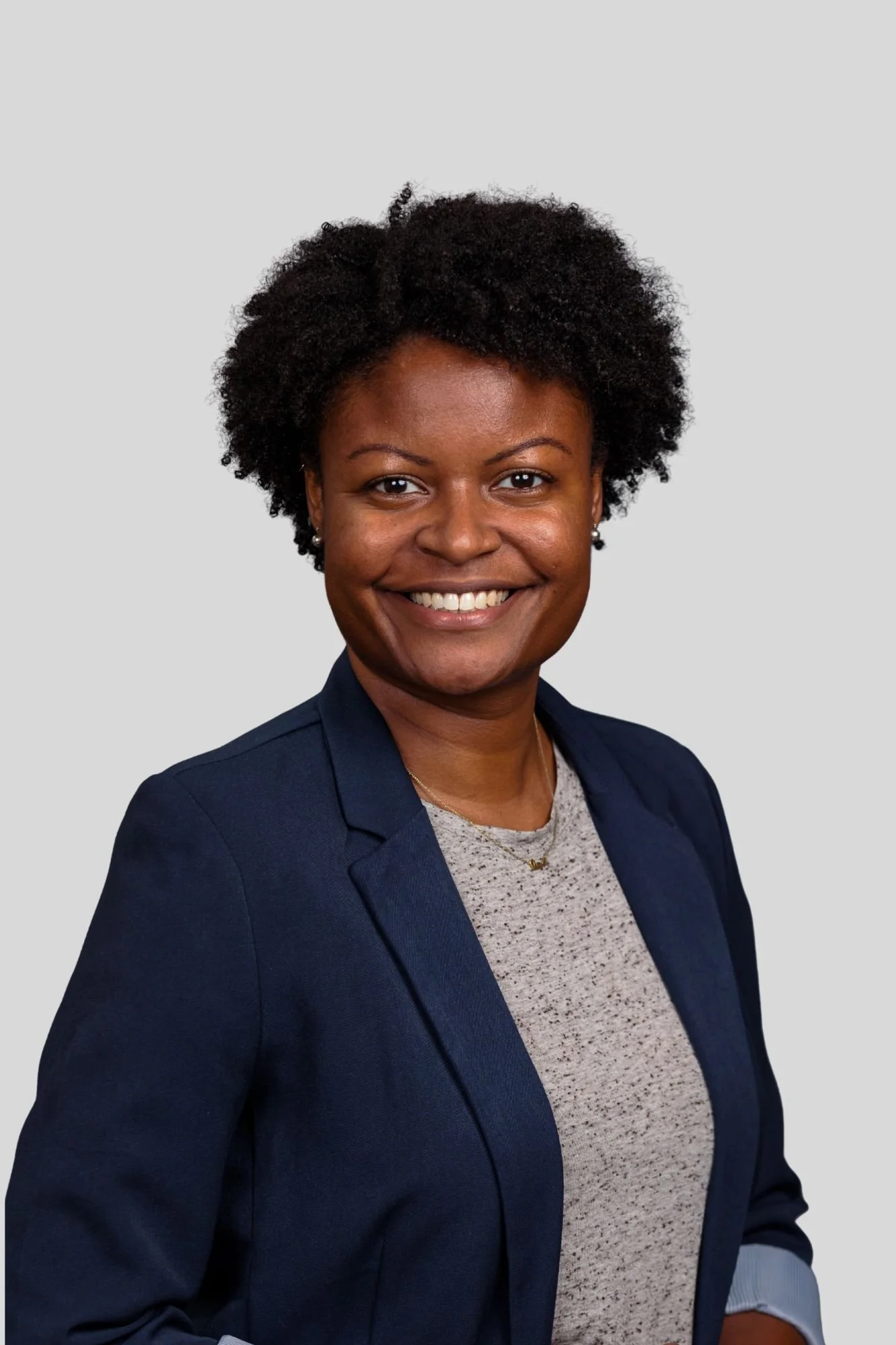 A headshot of an African American woman smiling, wearing a navy blazer over a patterned shirt, with a simple gray background.