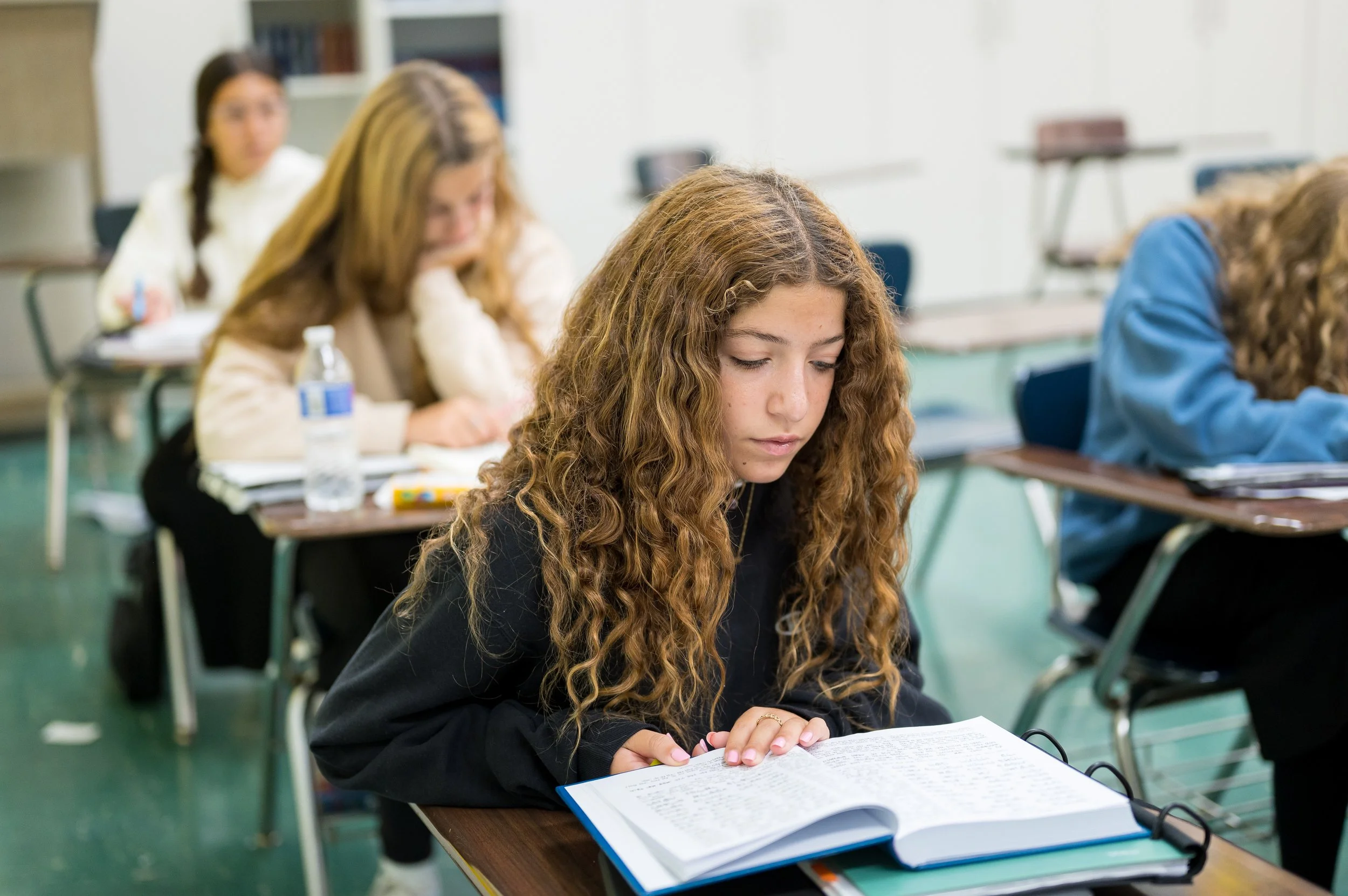 A young woman with long curly hair is sitting at a desk in a classroom, reading a book. Other students are visible in the background, also seated at desks, engaged in studying or writing.