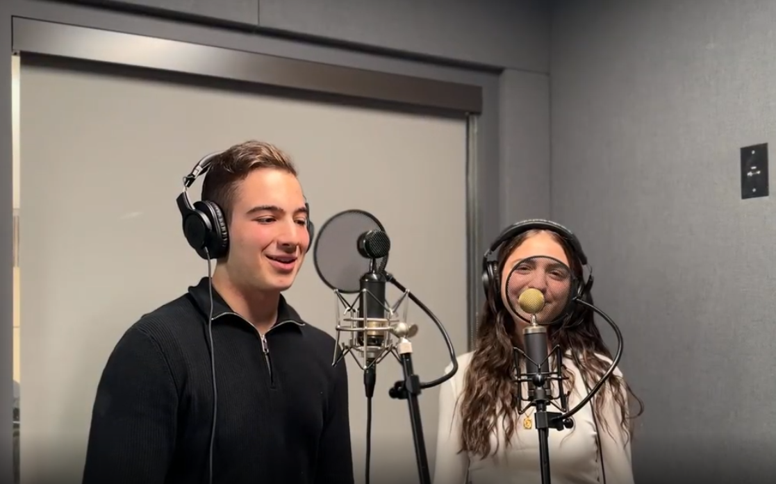 A young man and woman are recording vocals in a studio, each standing in front of a microphone with headphones on.