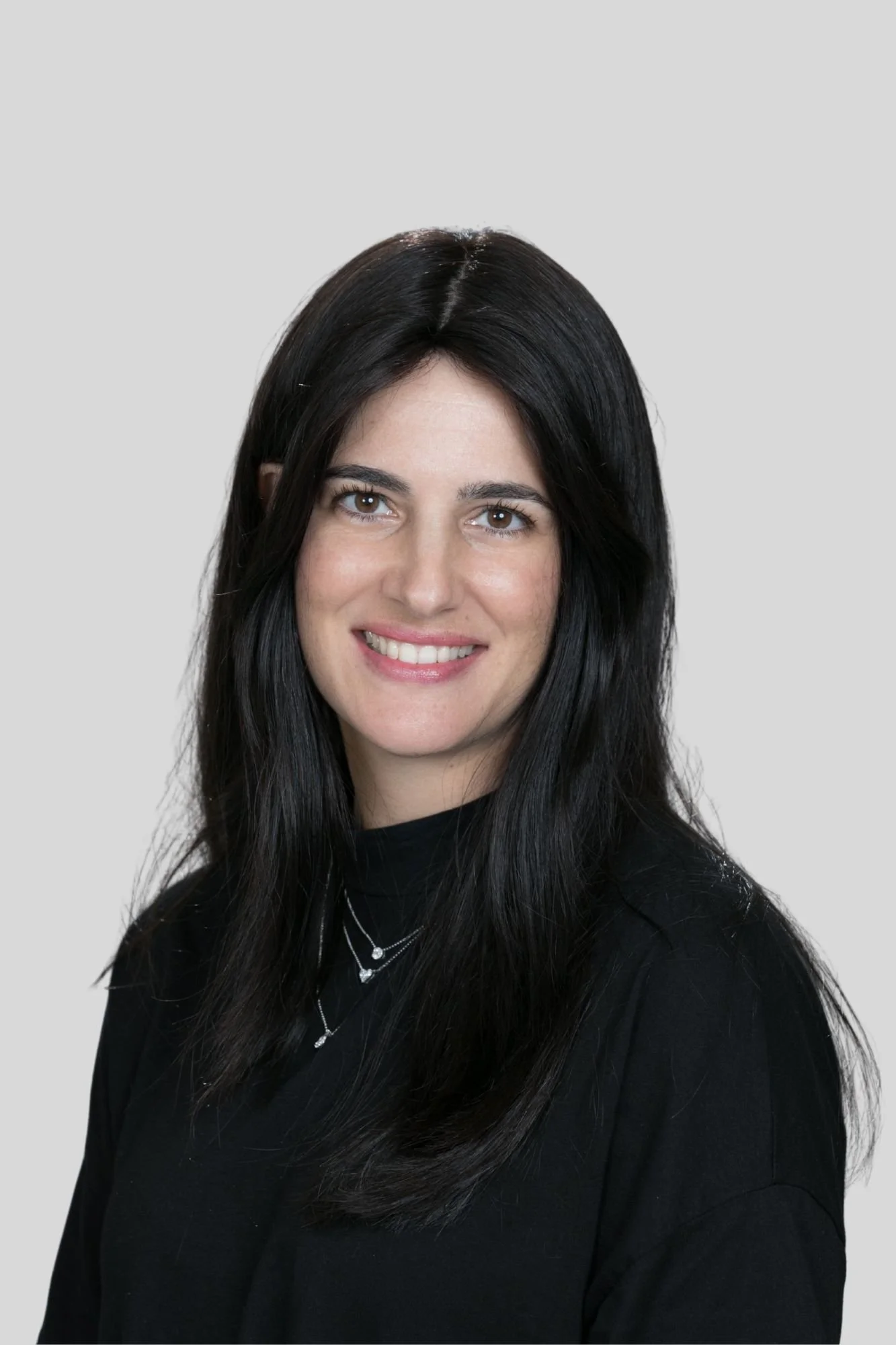 A woman with long black hair smiling, wearing a black top and layered necklaces, posed against a light gray background.