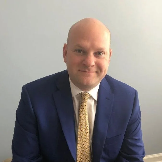 Bald man in a navy blue suit, white shirt, and yellow patterned tie sitting against a plain light gray background.