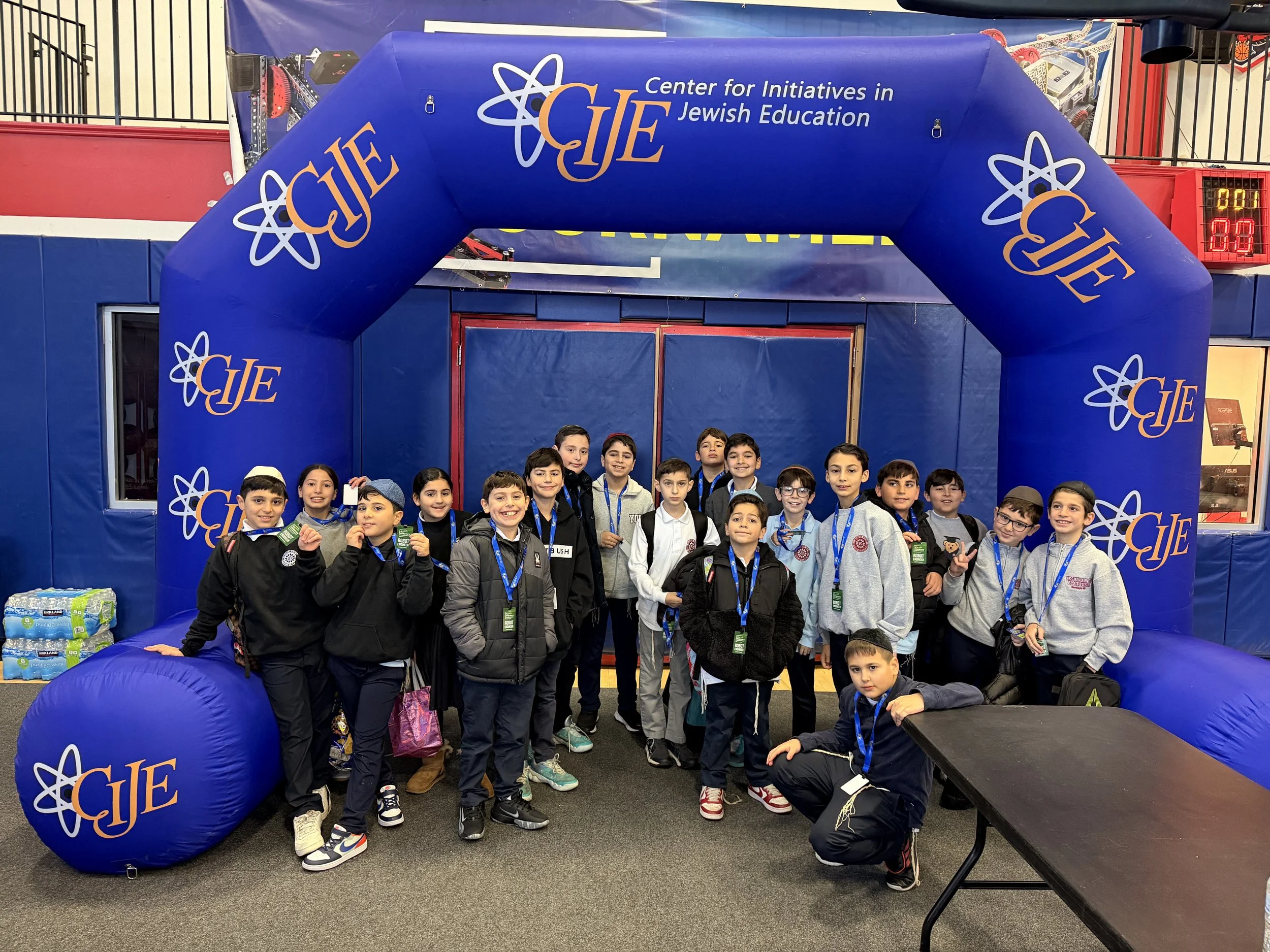 A group of children and teenagers standing inside an inflatable blue structure with the logo and text for the Center for Initiatives in Jewish Education (CIJE). The children are smiling and looking at the camera. Behind them, there is a partial view of a gym or event space.