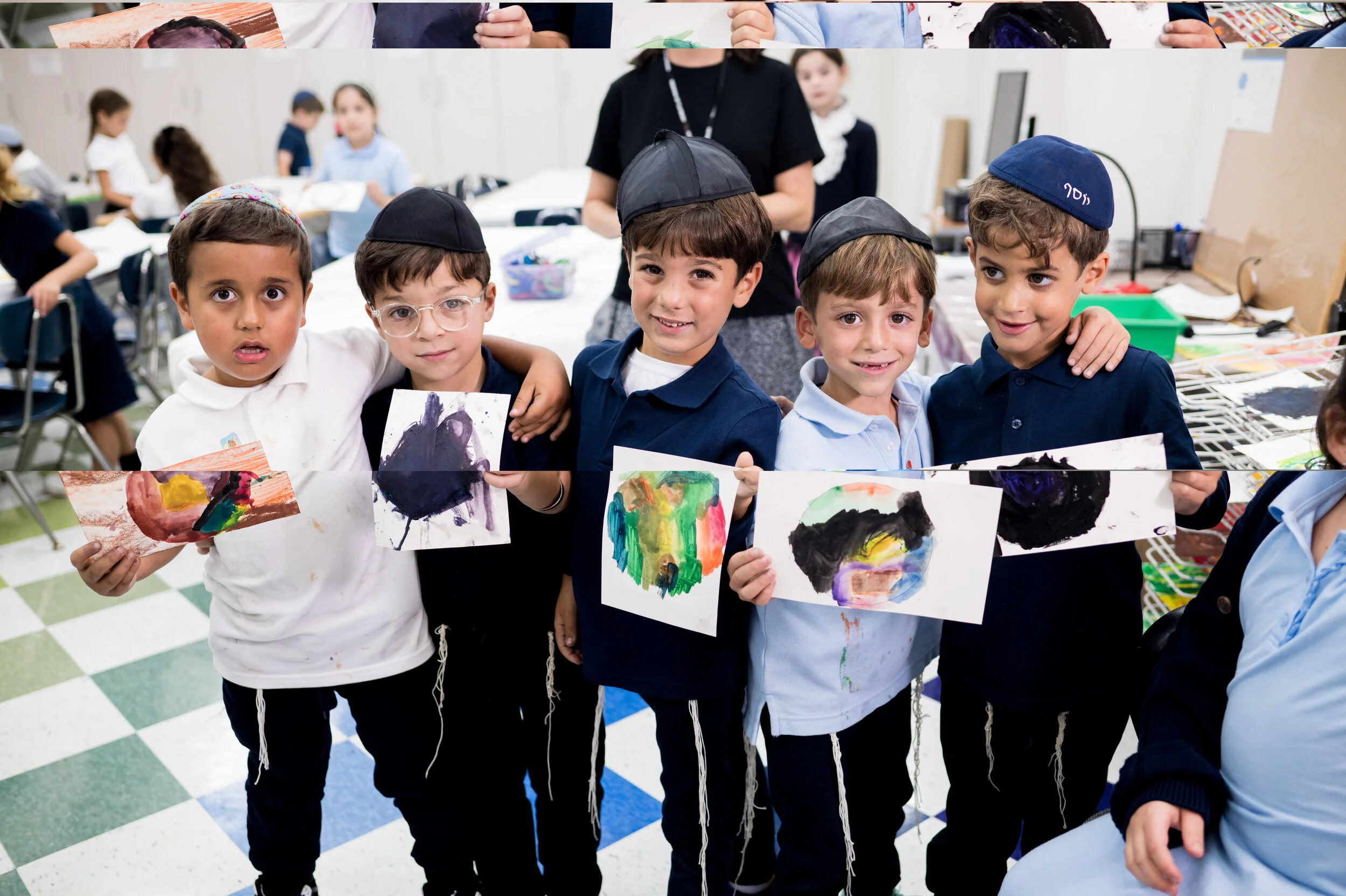 Five young boys standing together in a classroom, holding up artwork they created with abstract paintings, wearing kippahs, and smiling at the camera.