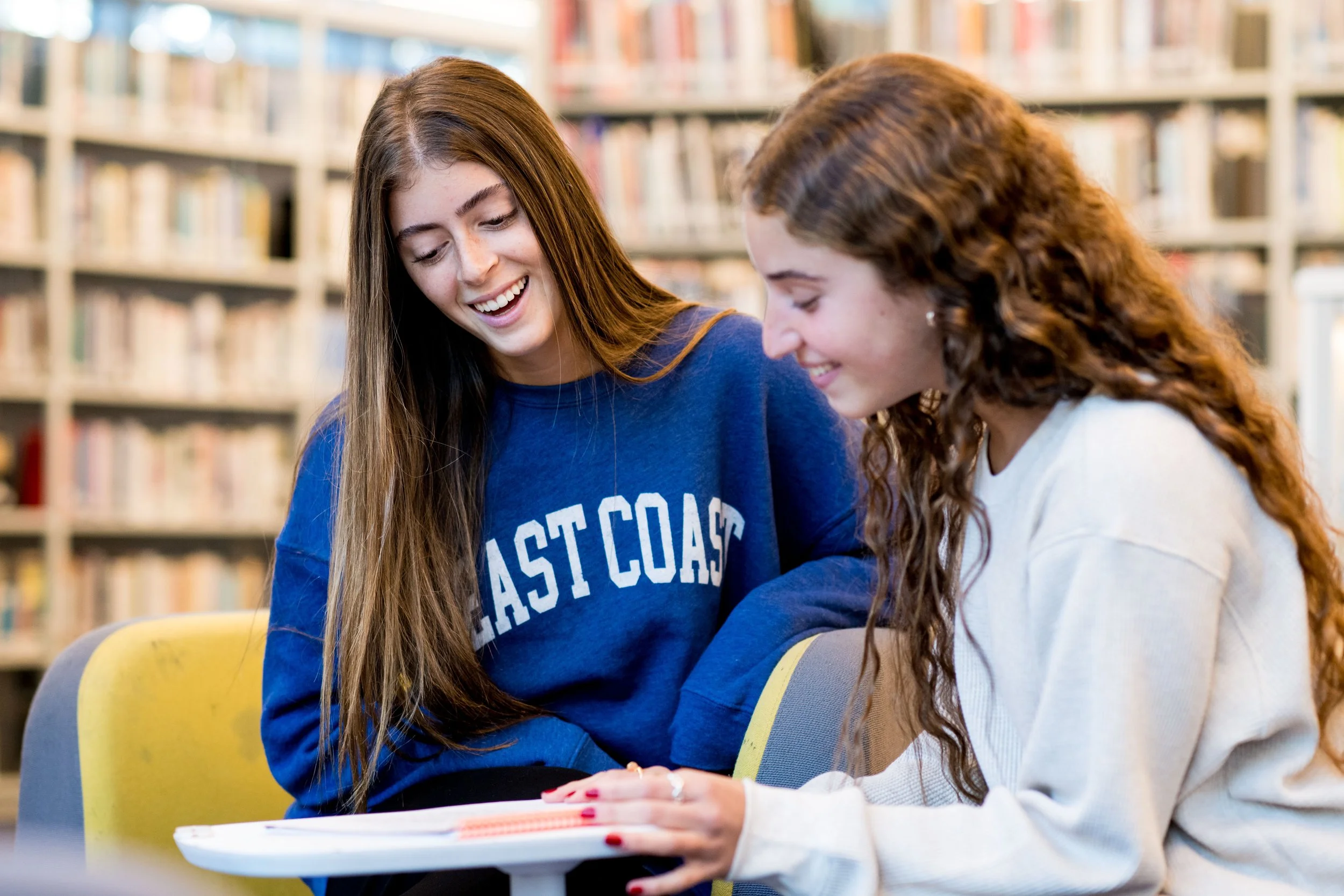 Two teenage girls are sitting at a table in a library, smiling and looking down at something on the table. Behind them are bookshelves filled with books.