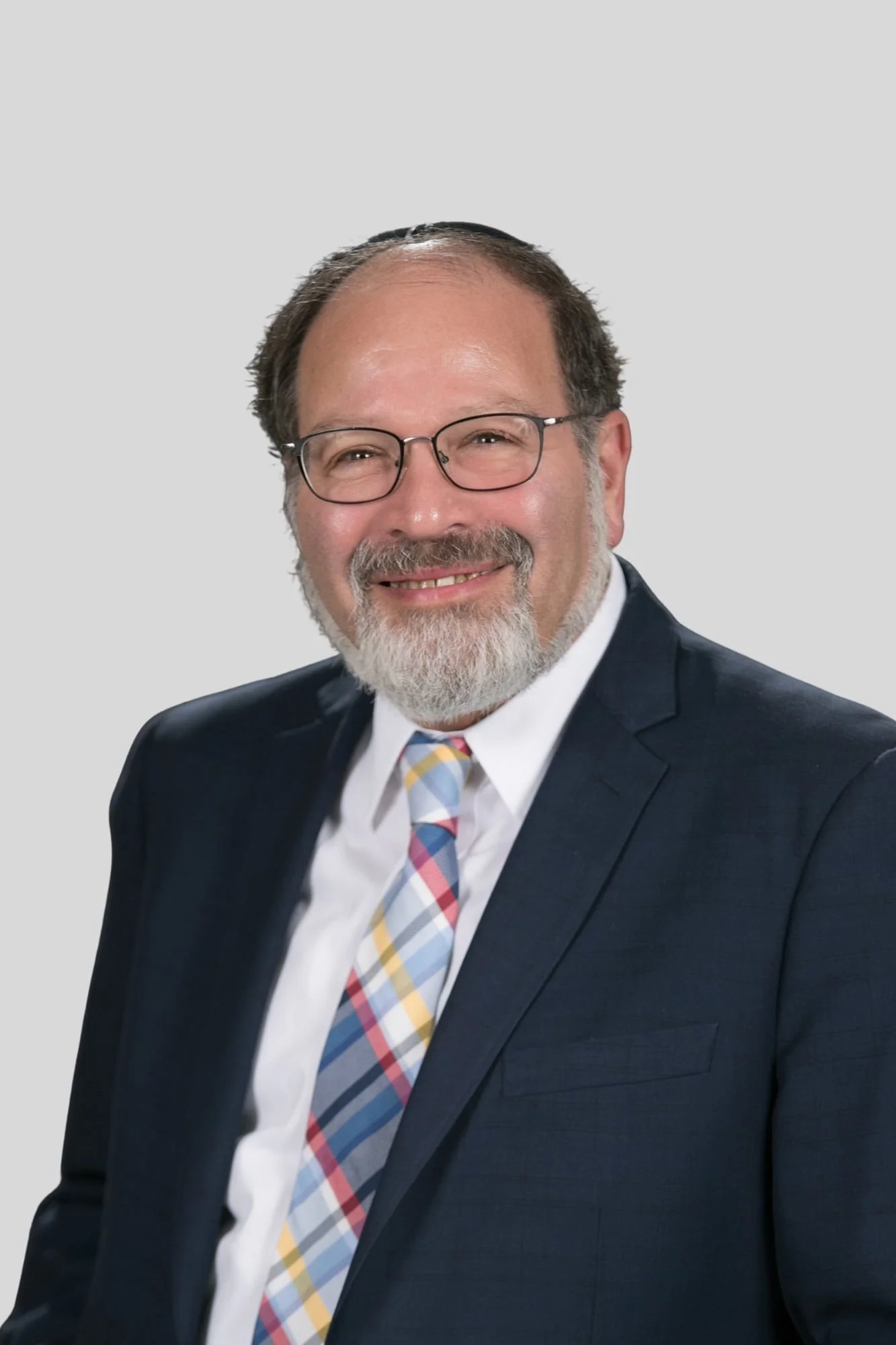 A middle-aged man in glasses, wearing a dark suit, white shirt, and a multicolored plaid tie, smiling against a plain light gray background.