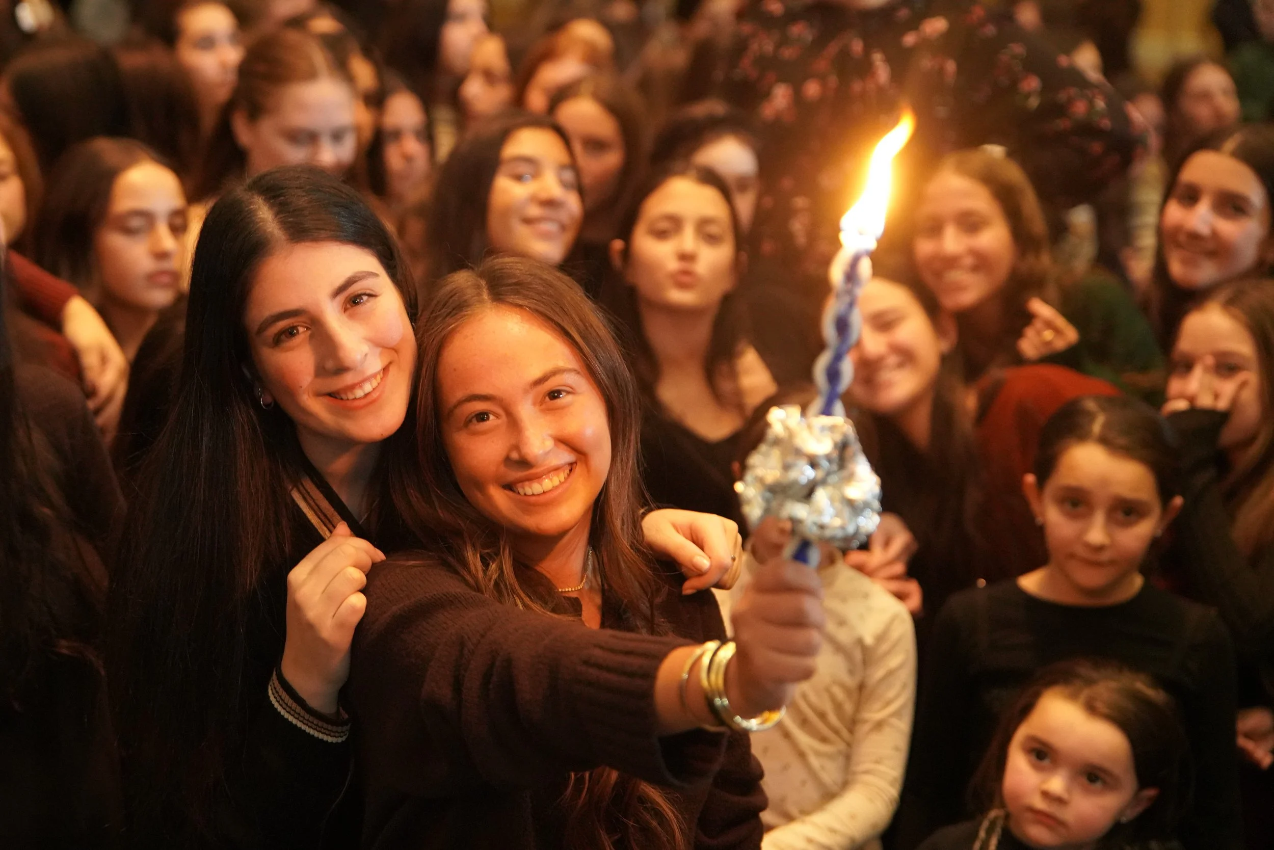 A group of women and children at a celebration, smiling and holding a lit menorah with a candle in the foreground.