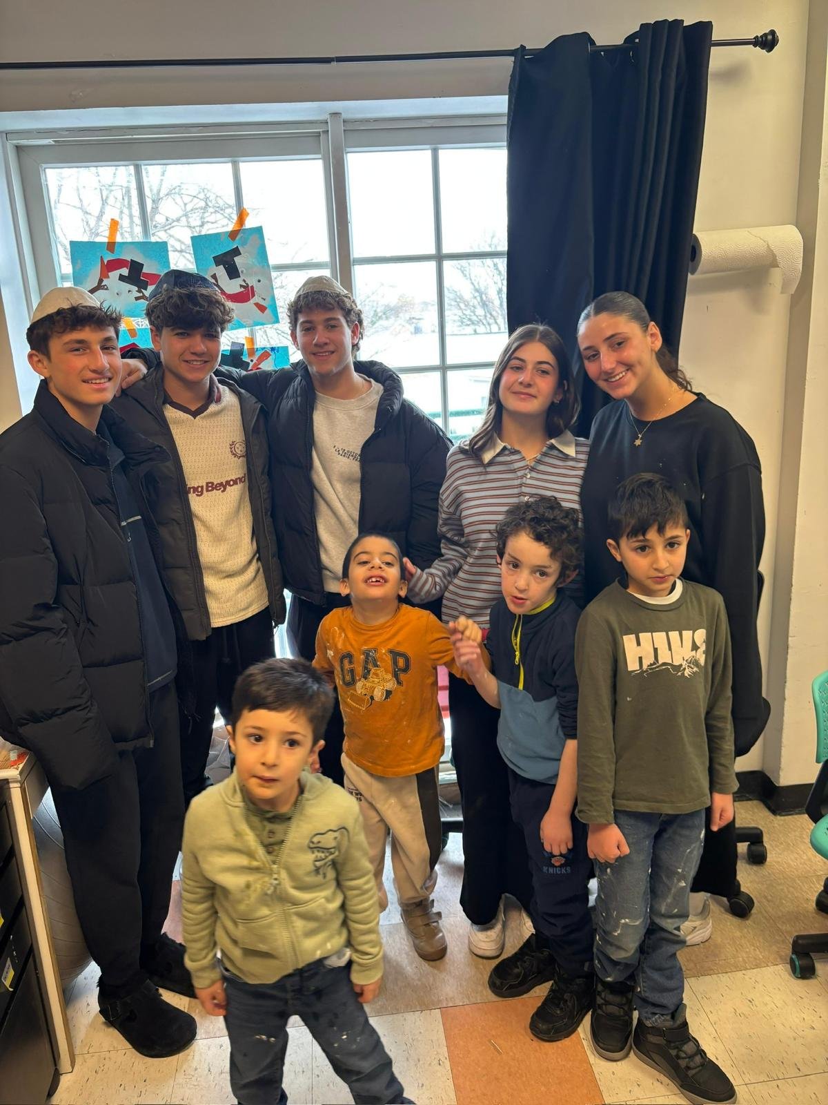 A group of children and teenagers posing together indoors near a large window with drawings hanging in the background, some smiling and making various gestures.