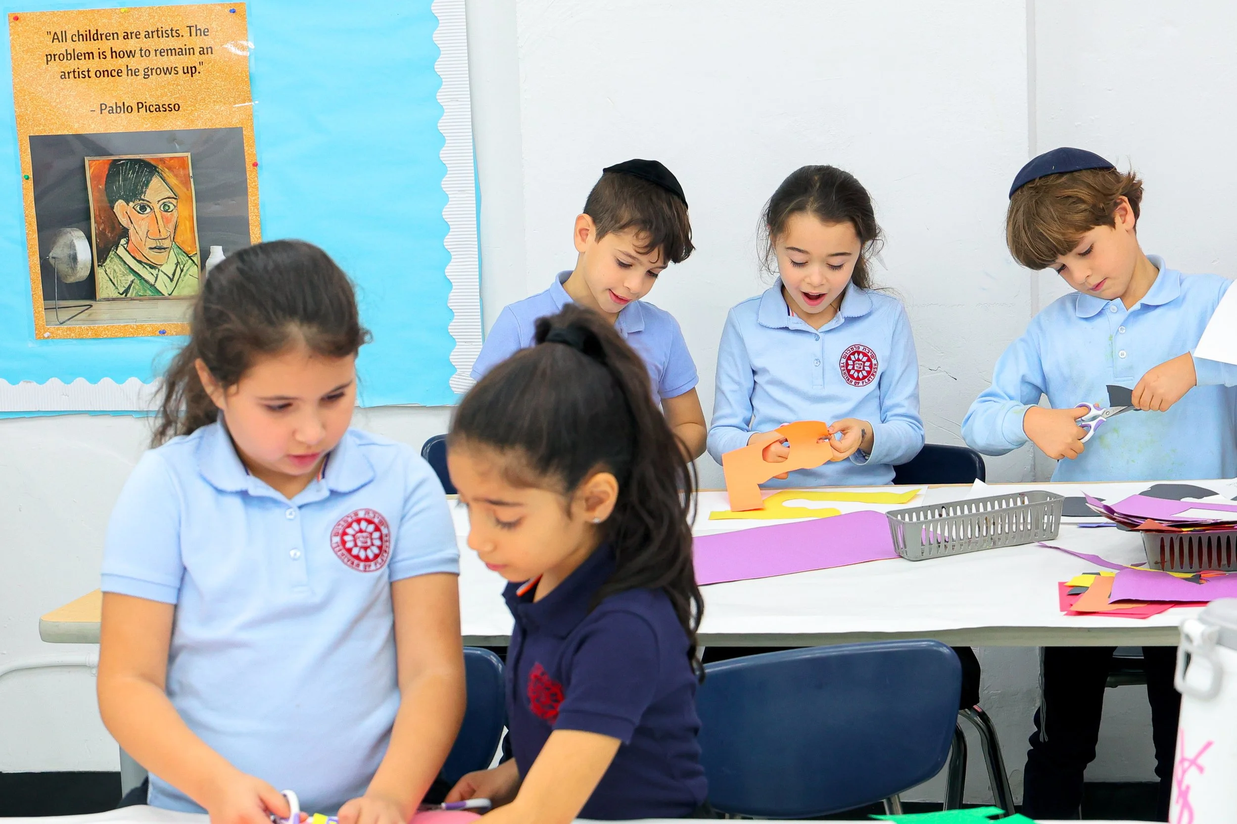 Children in school uniforms engaged in arts and crafts activities, sitting at tables with colored paper, scissors, and other craft supplies in a classroom setting. A bulletin board with a quote about children and art is visible in the background.