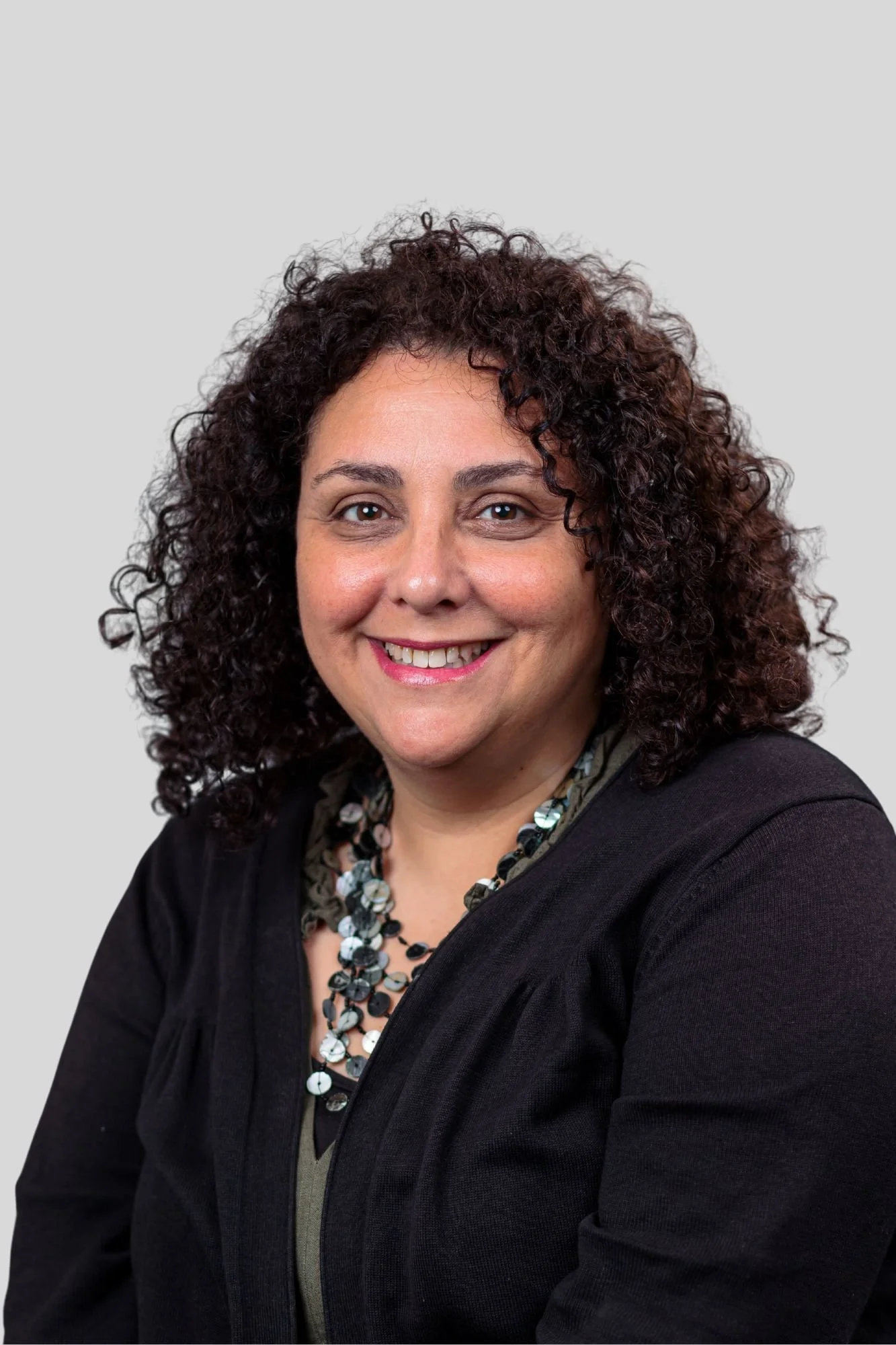 A woman with curly dark hair smiling, wearing a black blazer, necklace, and a patterned blouse, against a plain gray background.