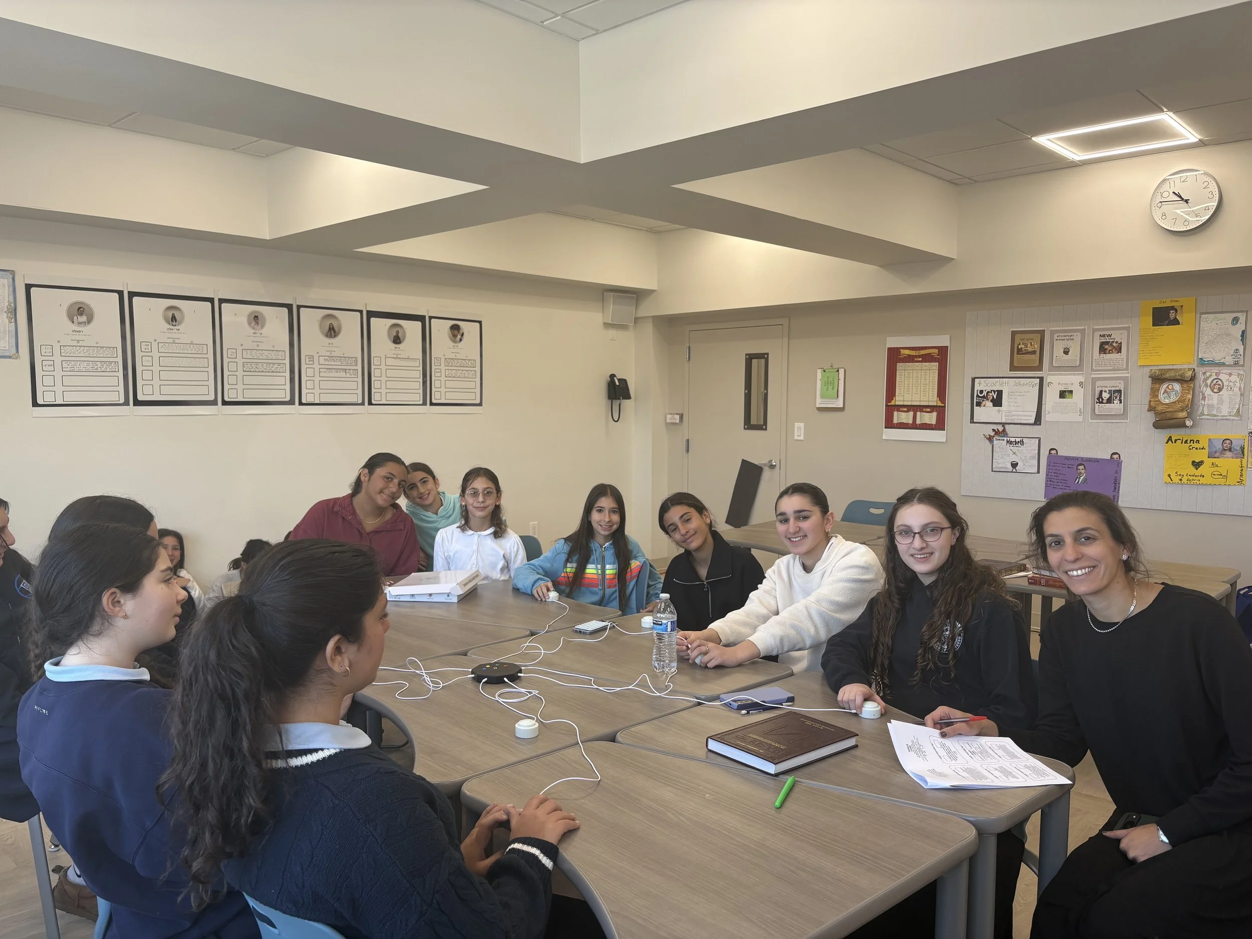Group of students sitting around a table in a classroom, smiling at the camera. The classroom has educational posters and a clock on the wall.
