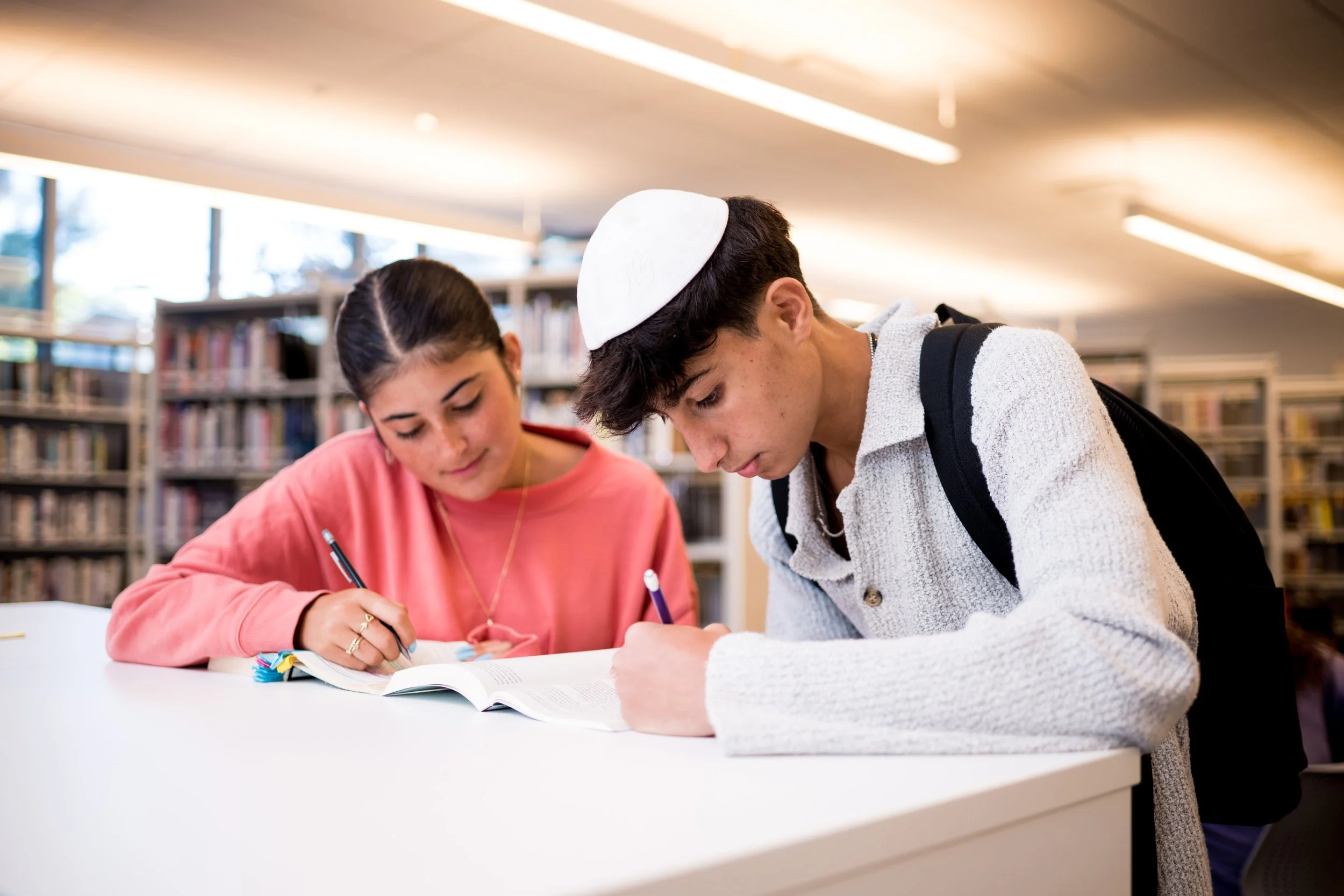 Two students, a girl and a boy, studying together at a library table with bookshelves in the background. The girl is writing in a notebook, and the boy is looking at an open book, wearing a yarmulke.
