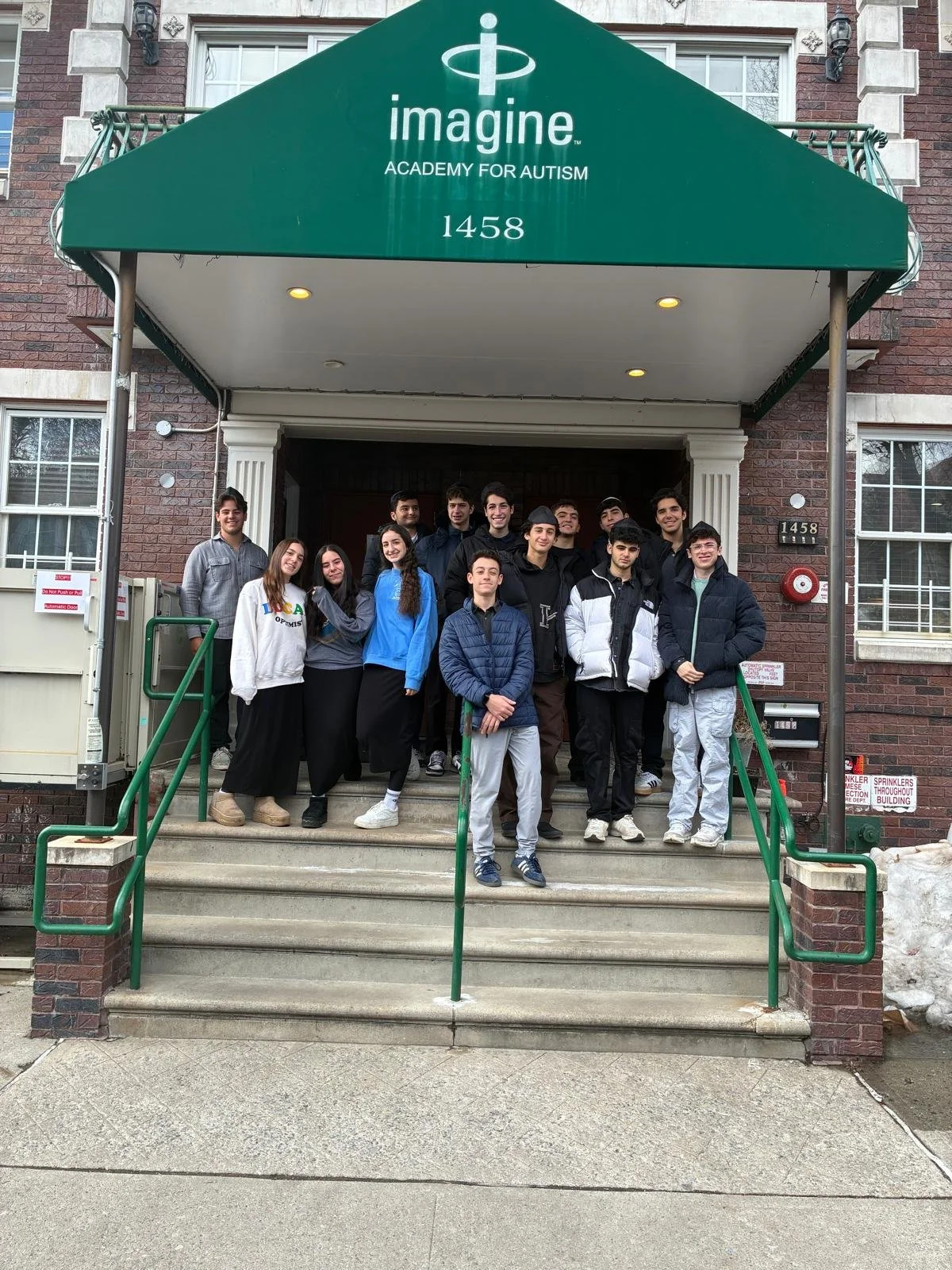 Group of teenagers standing on steps in front of a brick building with green awning, labeled 'imagine Academy for Autism,' and waving at the camera.