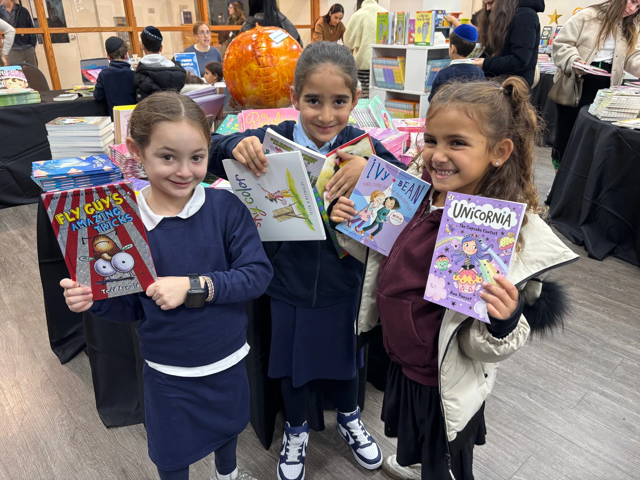 Three young girls smiling and holding colorful children's books at a bookstore or library event, surrounded by tables of books and other people in the background.