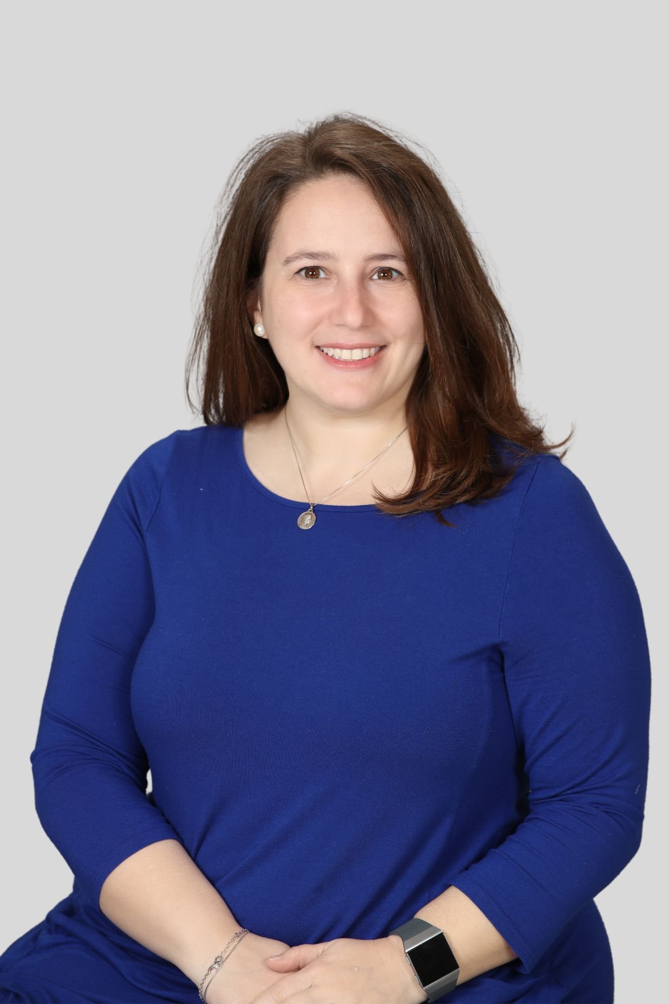 A woman with brown hair smiling, wearing a blue long-sleeved top, pearl earrings, a necklace, and a smartwatch, sitting against a plain light background.