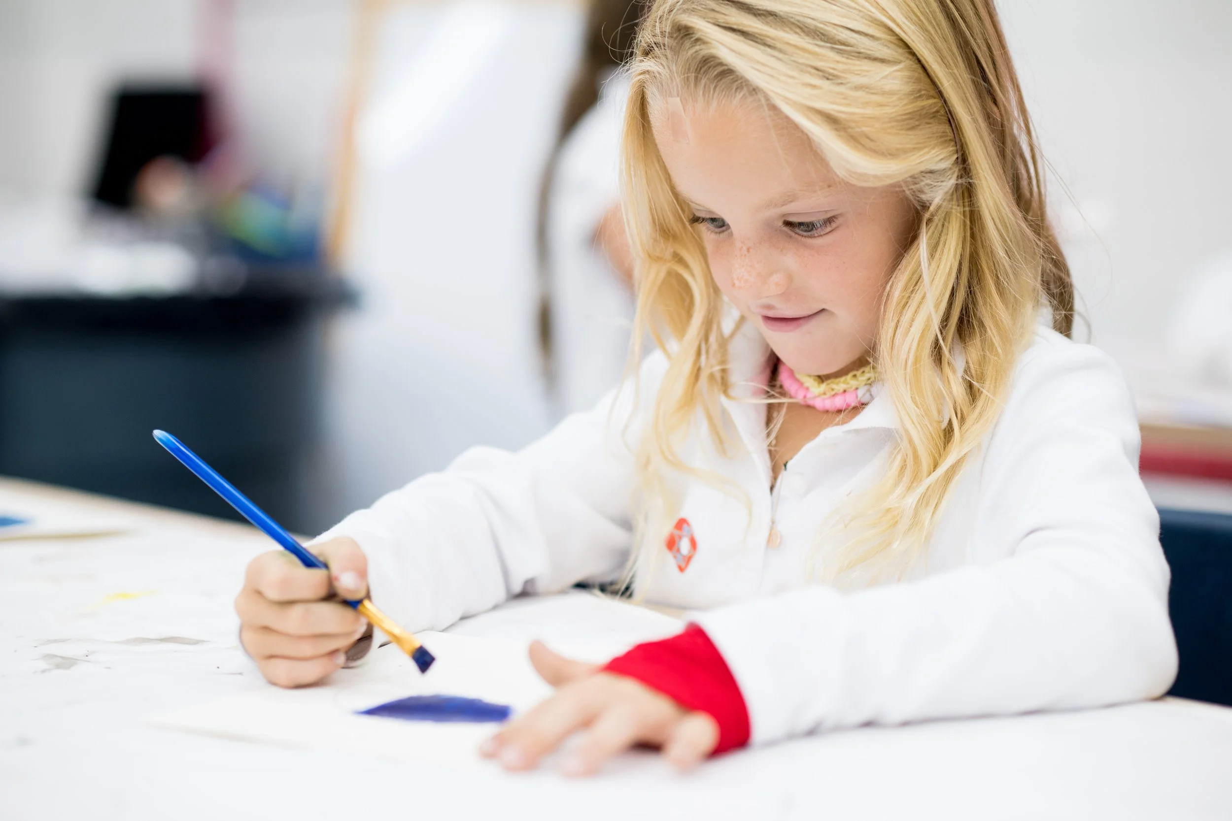A young girl with long blonde hair, freckles, and a pink necklace, is drawing with a blue pen on white paper at a desk.