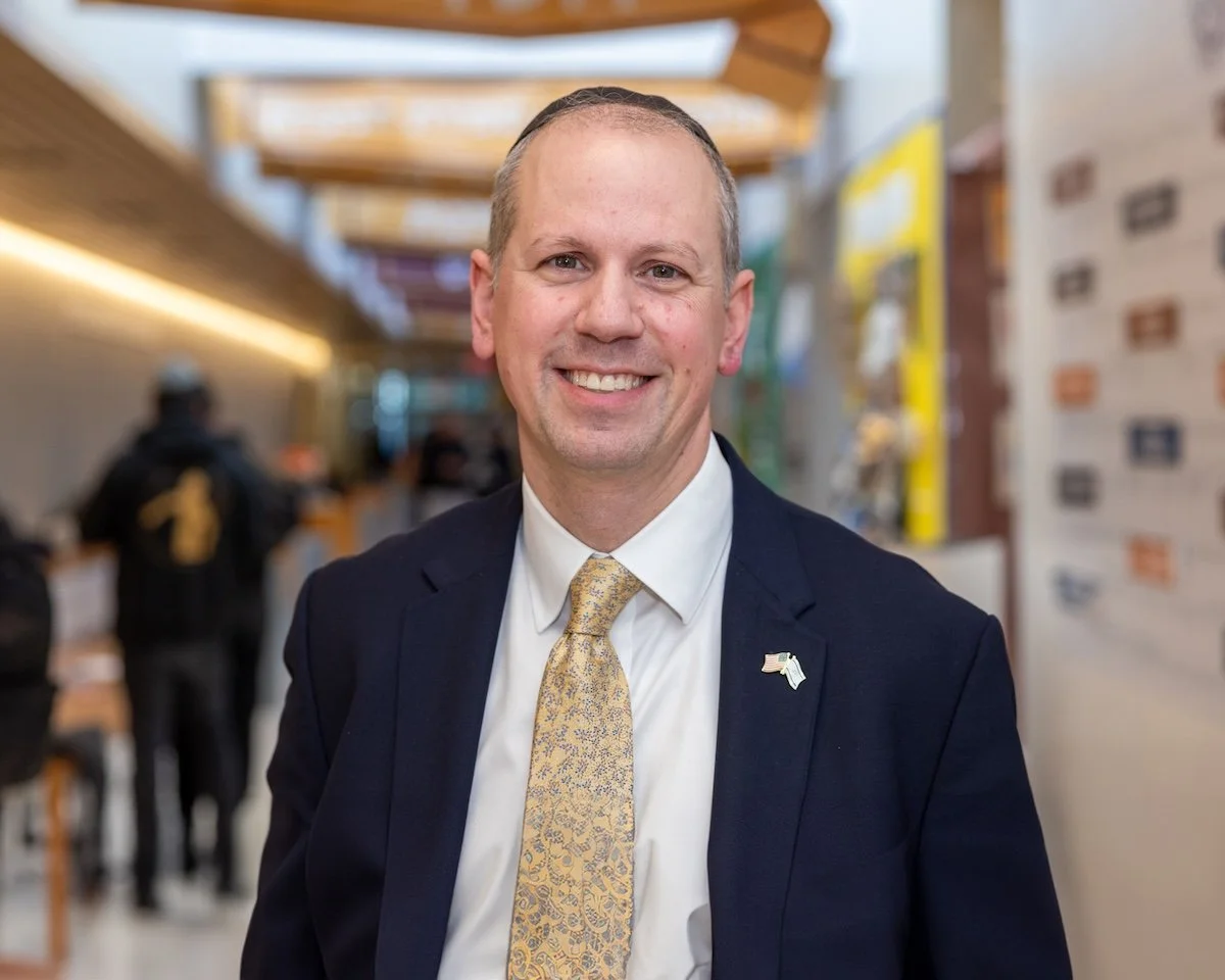 A man wearing a dark suit, white shirt, and yellow patterned tie standing and smiling in an indoor setting, with blurred background of people and colorful booths or displays.