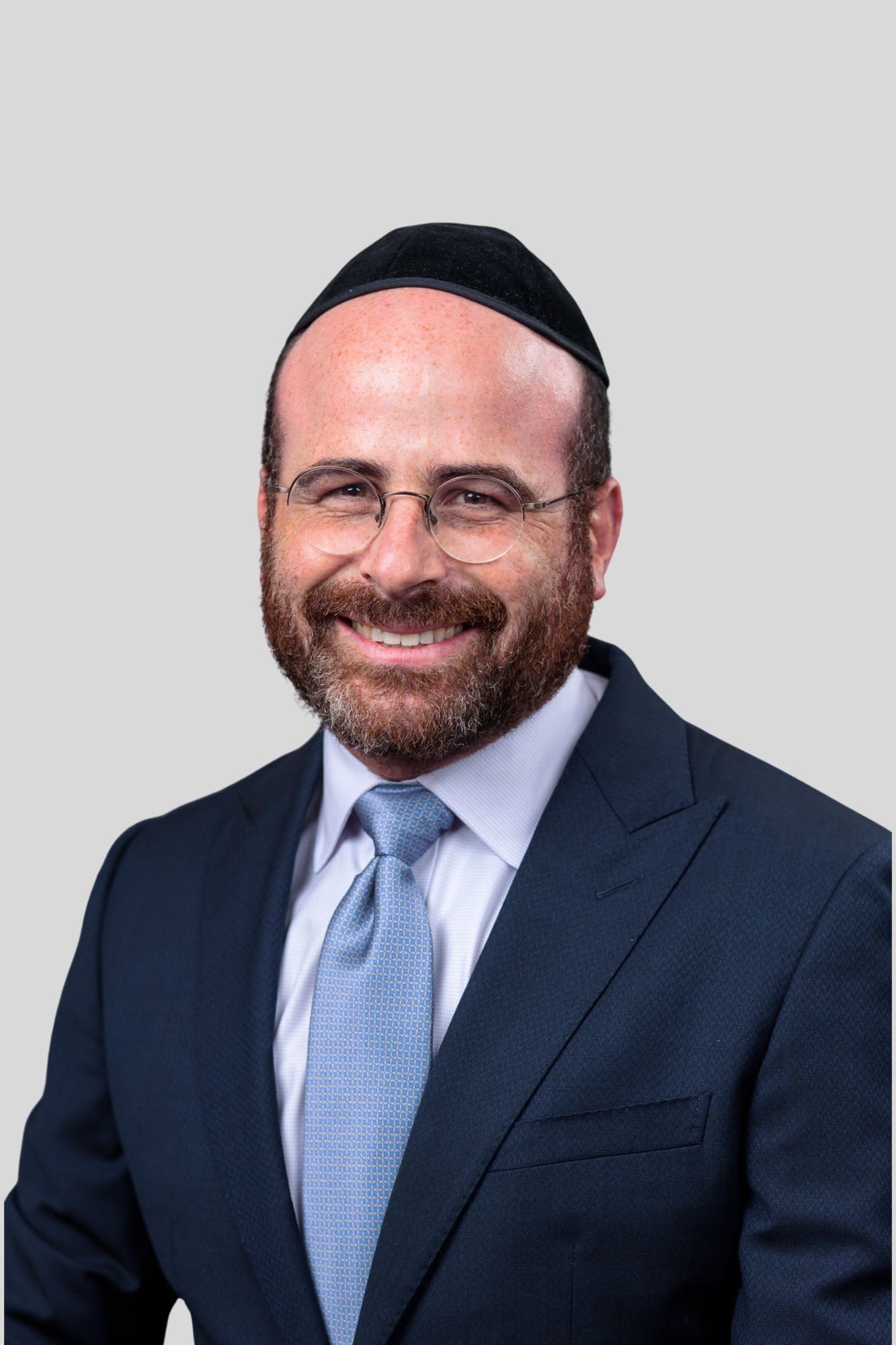 A man dressed in a dark blue suit, light blue tie, white shirt, wearing glasses and a black kippah, smiling against a plain light gray background.