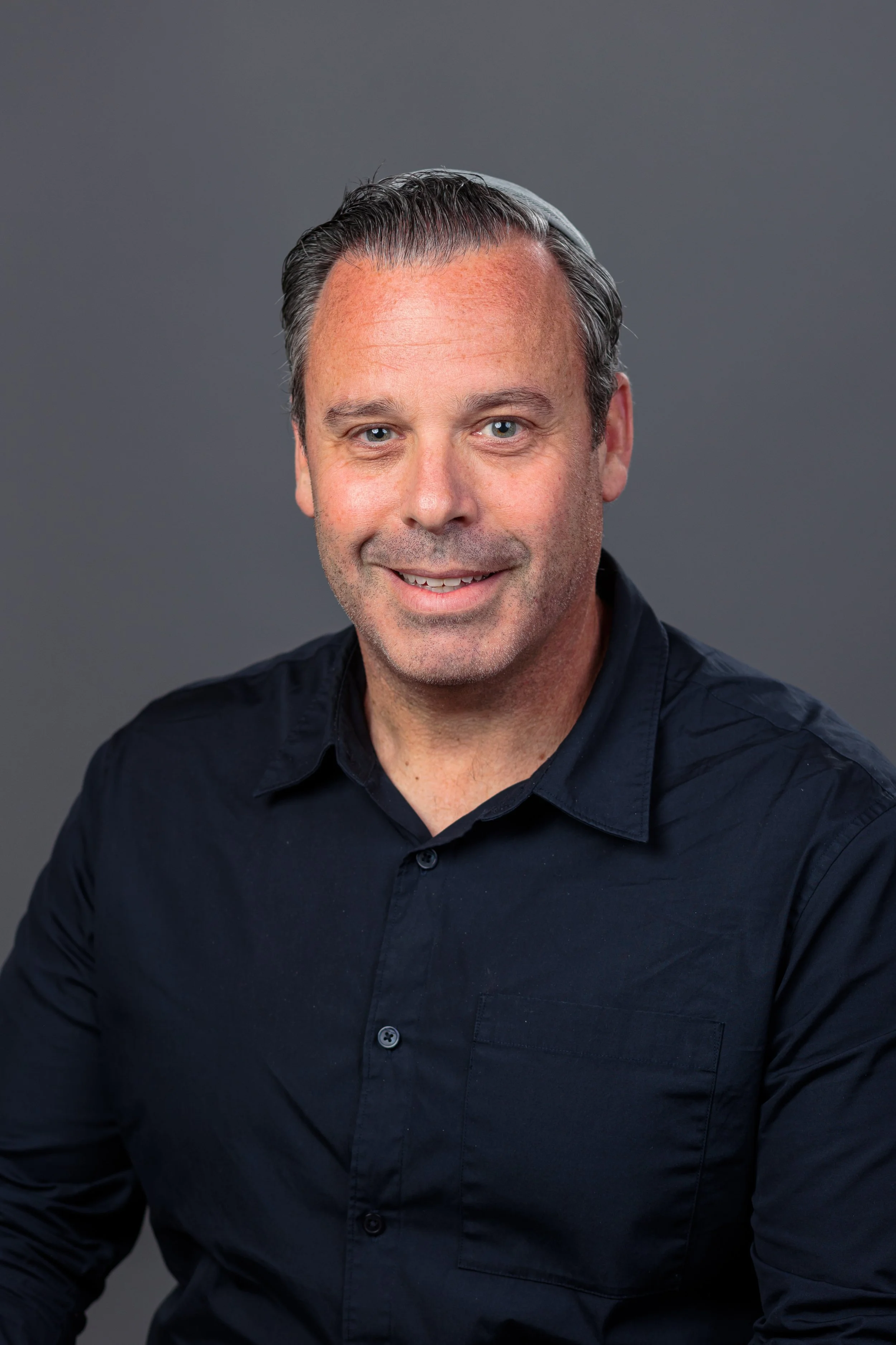 A portrait of a man with short, dark hair with some gray, wearing a dark navy blue button-up shirt, smiling against a plain gray background.
