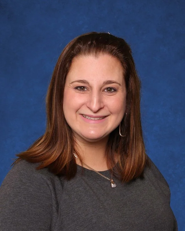 A woman with shoulder-length brown hair, smiling, wearing a dark gray top and a silver necklace, posed against a plain blue background.