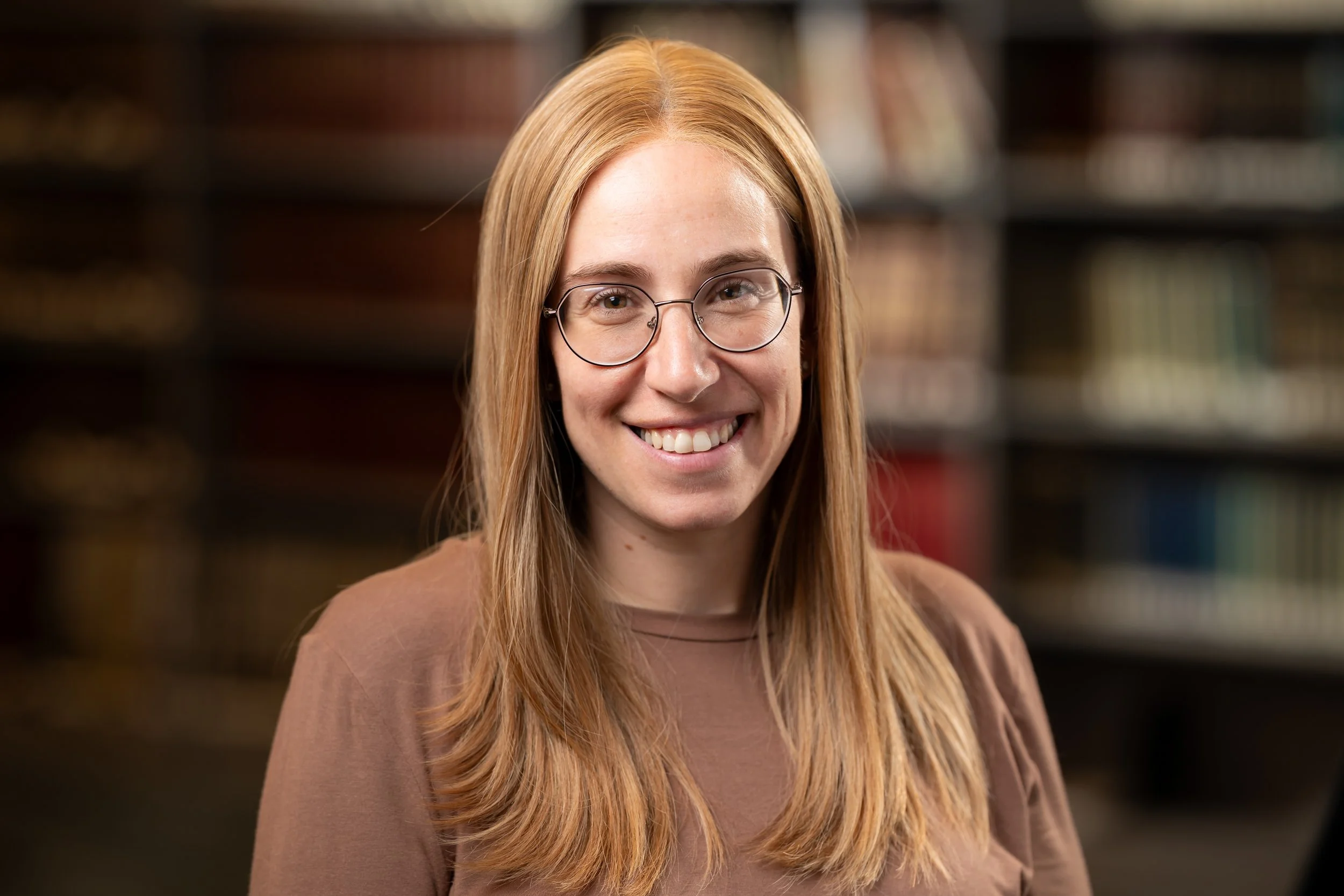 A young woman with long red hair, wearing glasses, smiling, standing in front of a background of bookshelves.