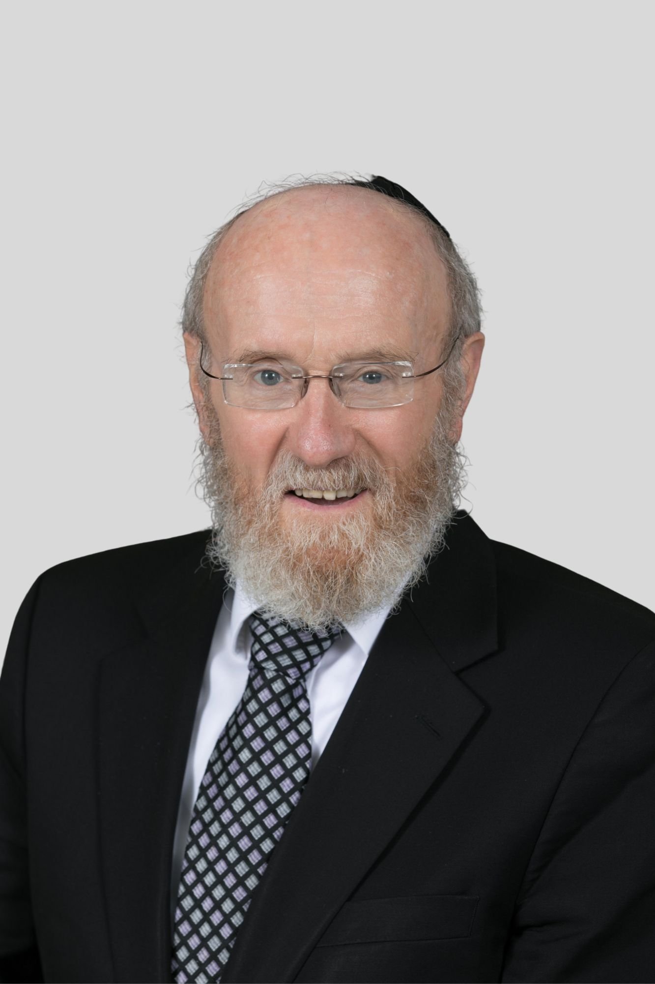 A middle-aged man with glasses, a beard, and a mustache, wearing a black suit, white shirt, and patterned tie, smiling against a plain light grey background.