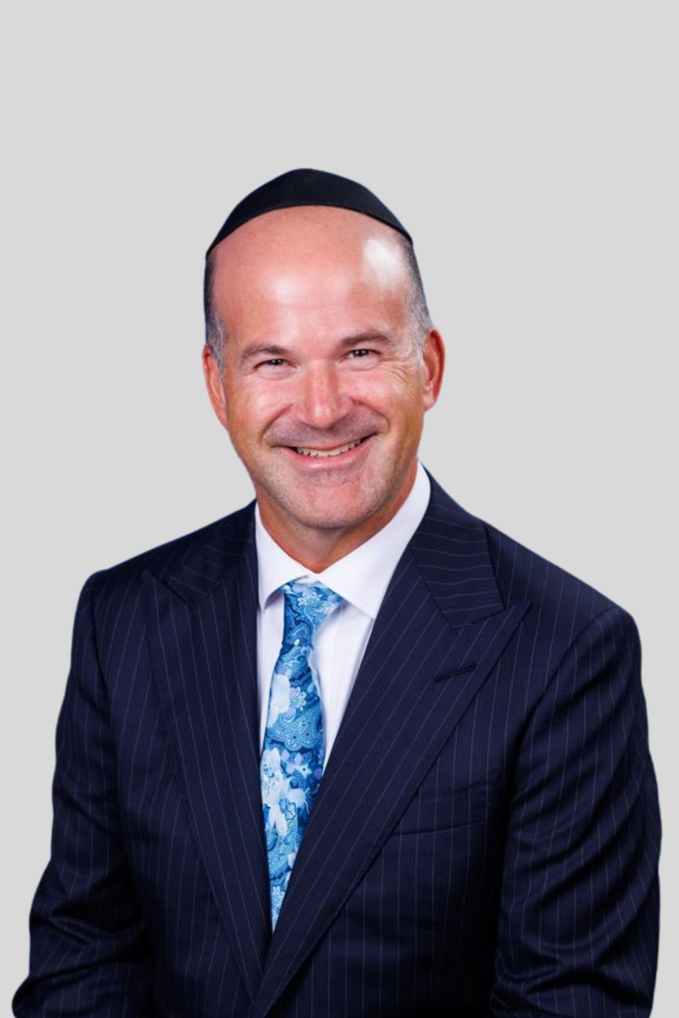 A smiling man wearing a dark pinstripe suit, a white shirt, a blue paisley tie, and a black kippah, posed against a light gray background.