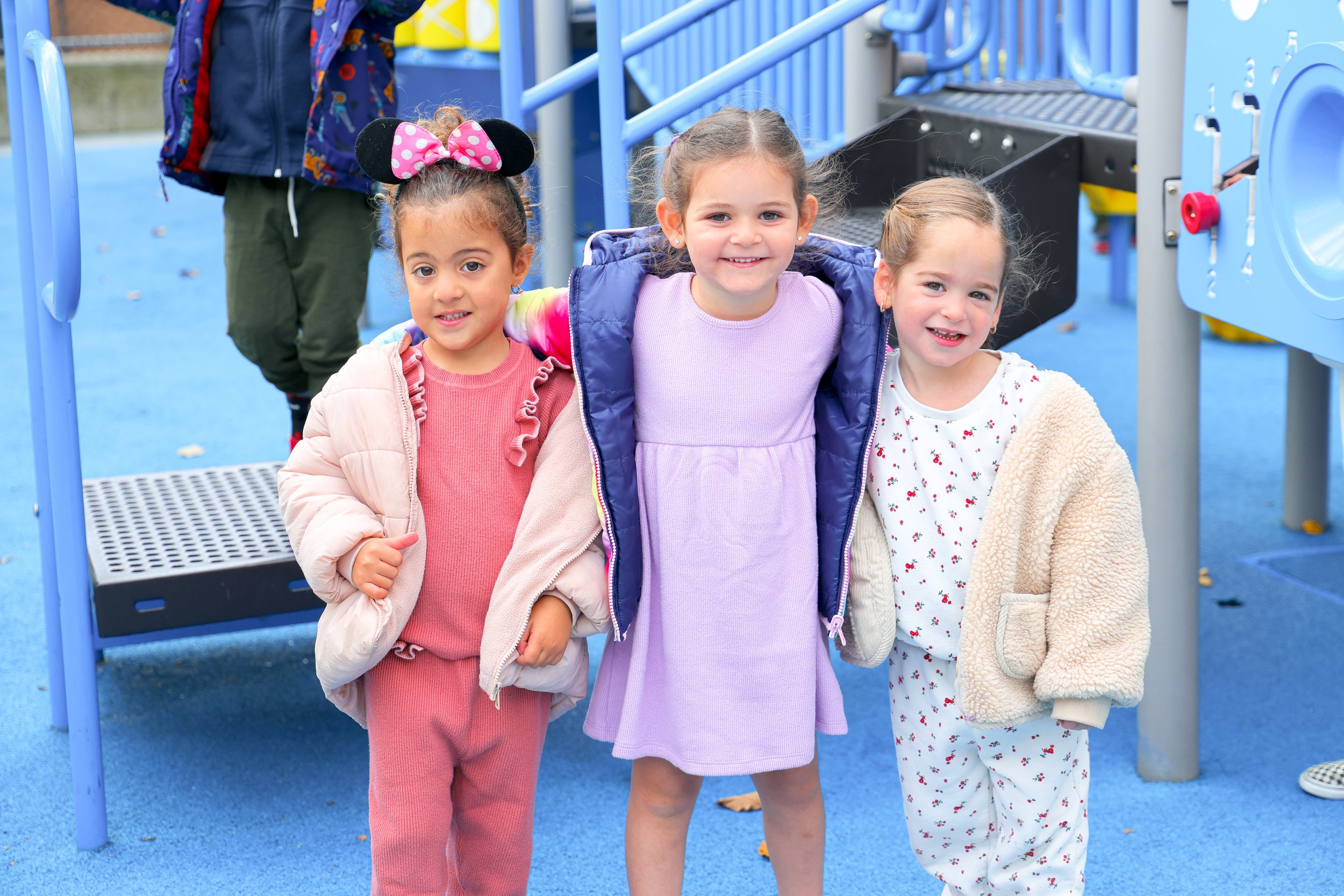 Three young girls standing closely together on a playground, smiling at the camera. Two girls are wearing pink and cream outfits, and the girl in the middle is in a purple dress with a blue jacket. Behind them, there is a blue playground structure.