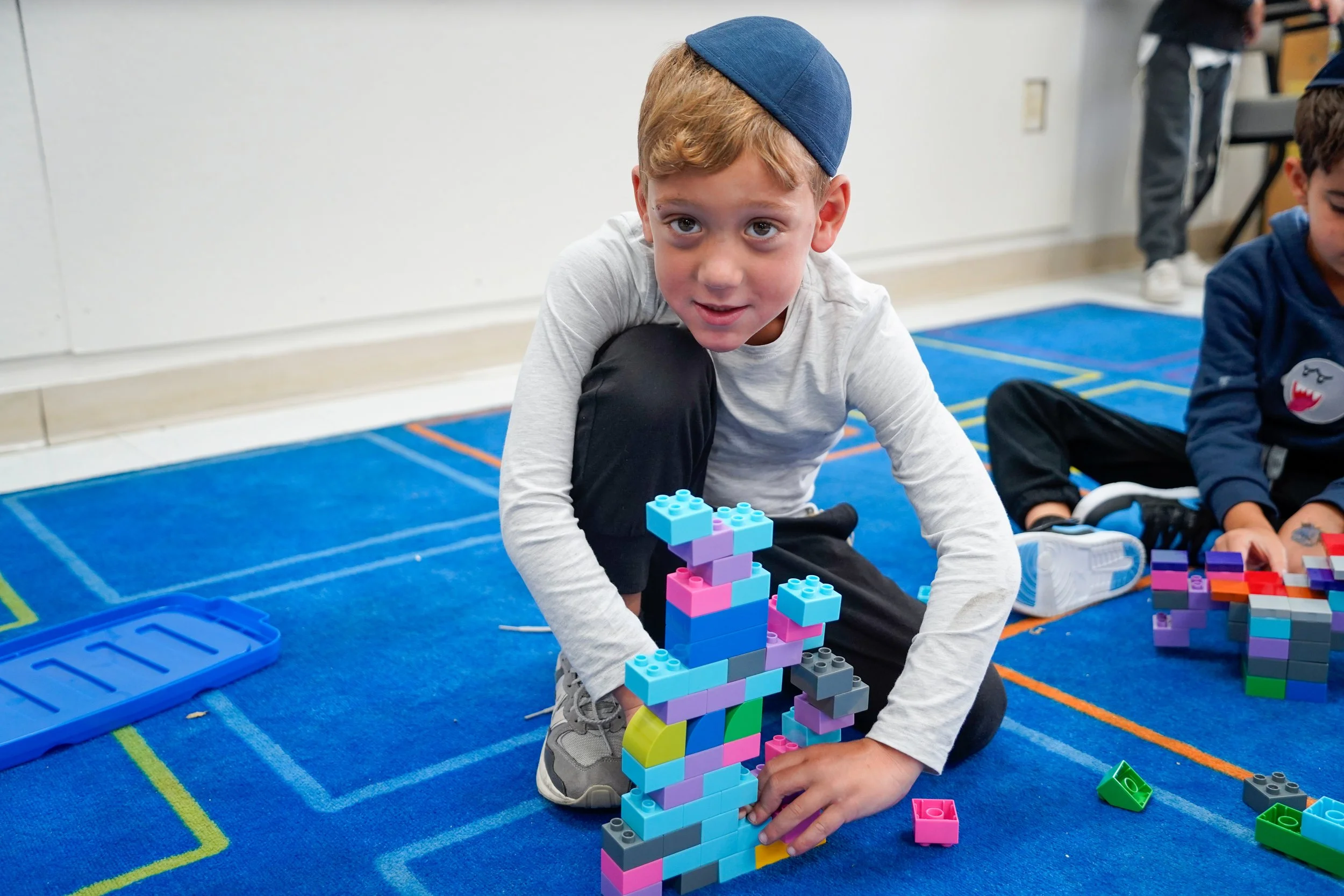A young boy with red hair, wearing a blue kippah, white long sleeve shirt, and gray sneakers, is playing with colorful plastic building blocks on a blue carpeted floor in a classroom. Other children and classroom furniture are visible in the background.