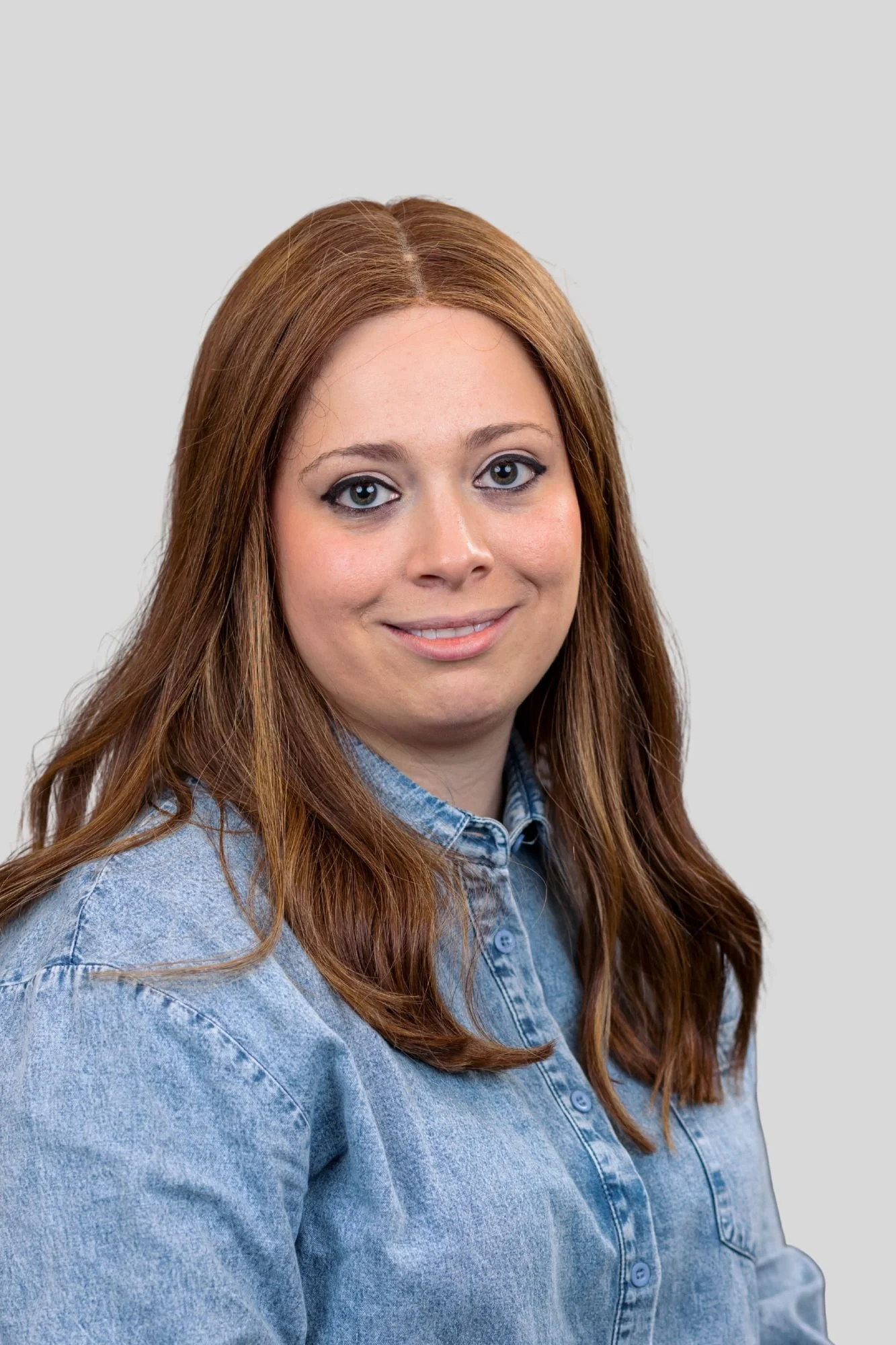 A woman with long red hair wearing a denim jacket against a plain gray background.