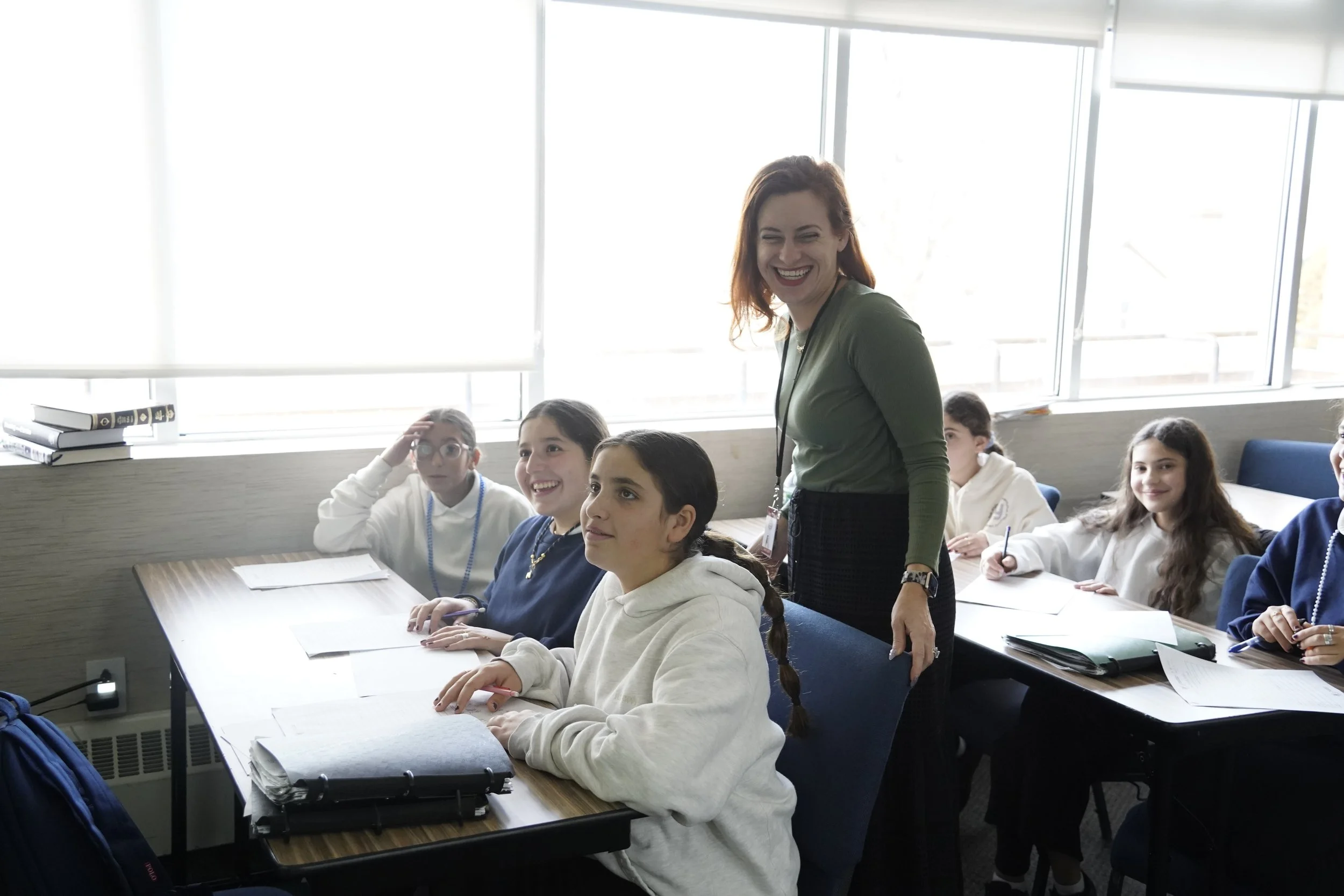A classroom with a group of students seated at desks and a teacher standing among them, smiling. The students appear engaged, some with notebooks and pens. Large windows in the background allow natural light to illuminate the room.